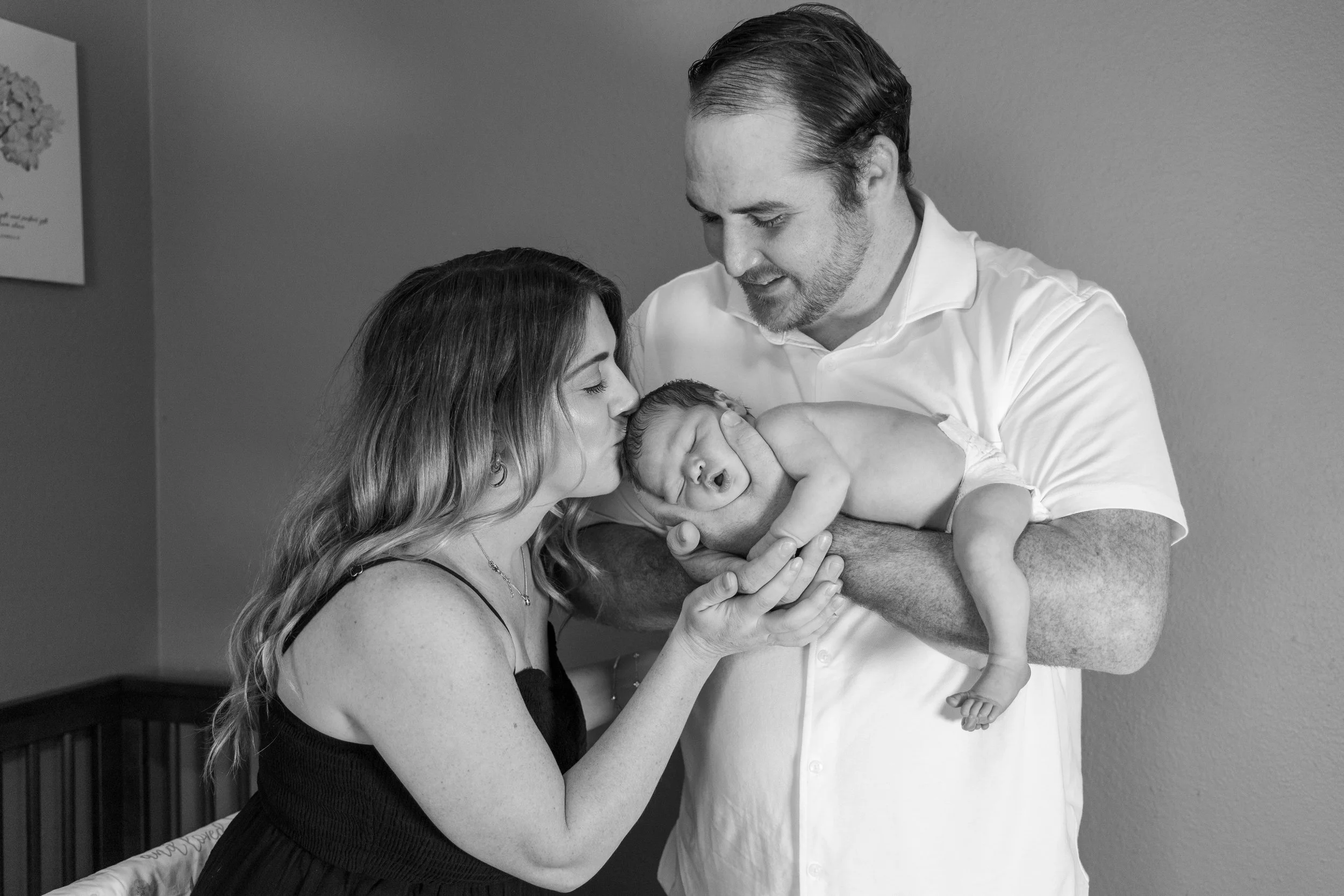 A woman is holding a newborn baby close to her face, kissing the baby. A man, possibly the father, is supporting the baby with both hands and looking down at the child. The scene appears to be a tender moment in a hospital or home setting.