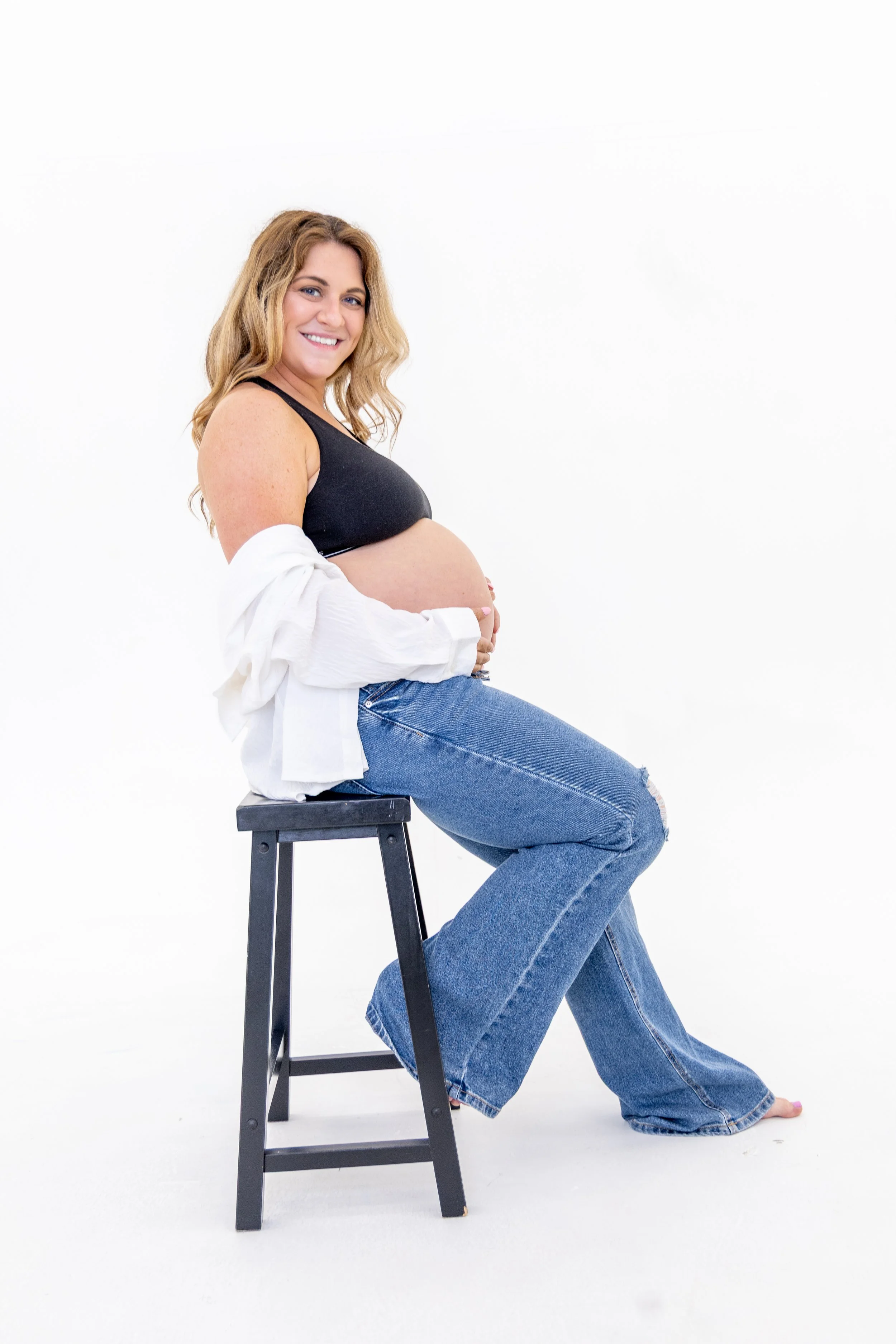 A pregnant woman with blonde hair sitting on a black stool, smiling, wearing a black tank top, a white shirt hanging off her shoulders, and ripped blue jeans, against a white background.