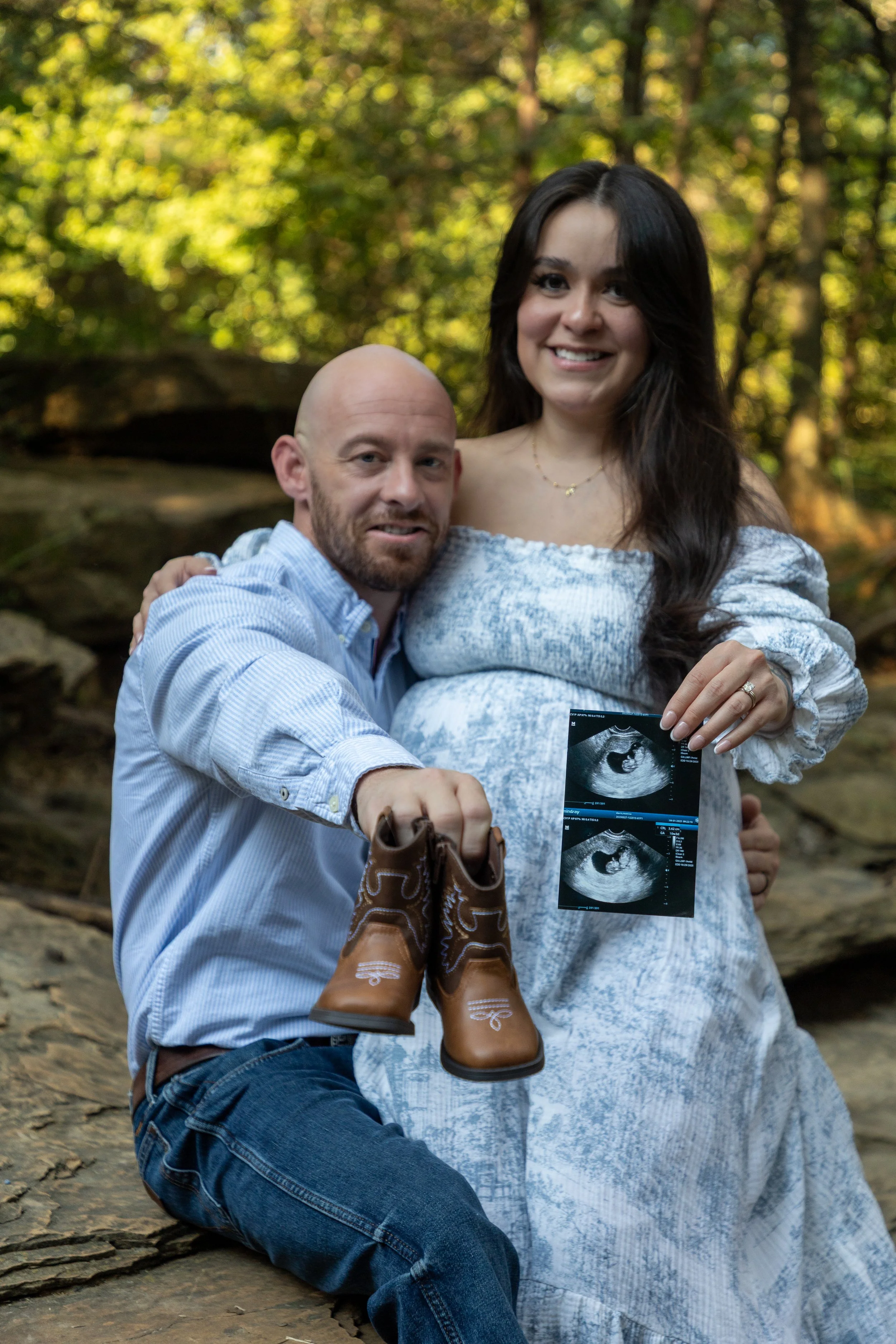 A man and a pregnant woman sitting outdoors in a wooded area, holding an ultrasound image of a developing baby and a pair of tiny brown baby shoes, celebrating pregnancy.