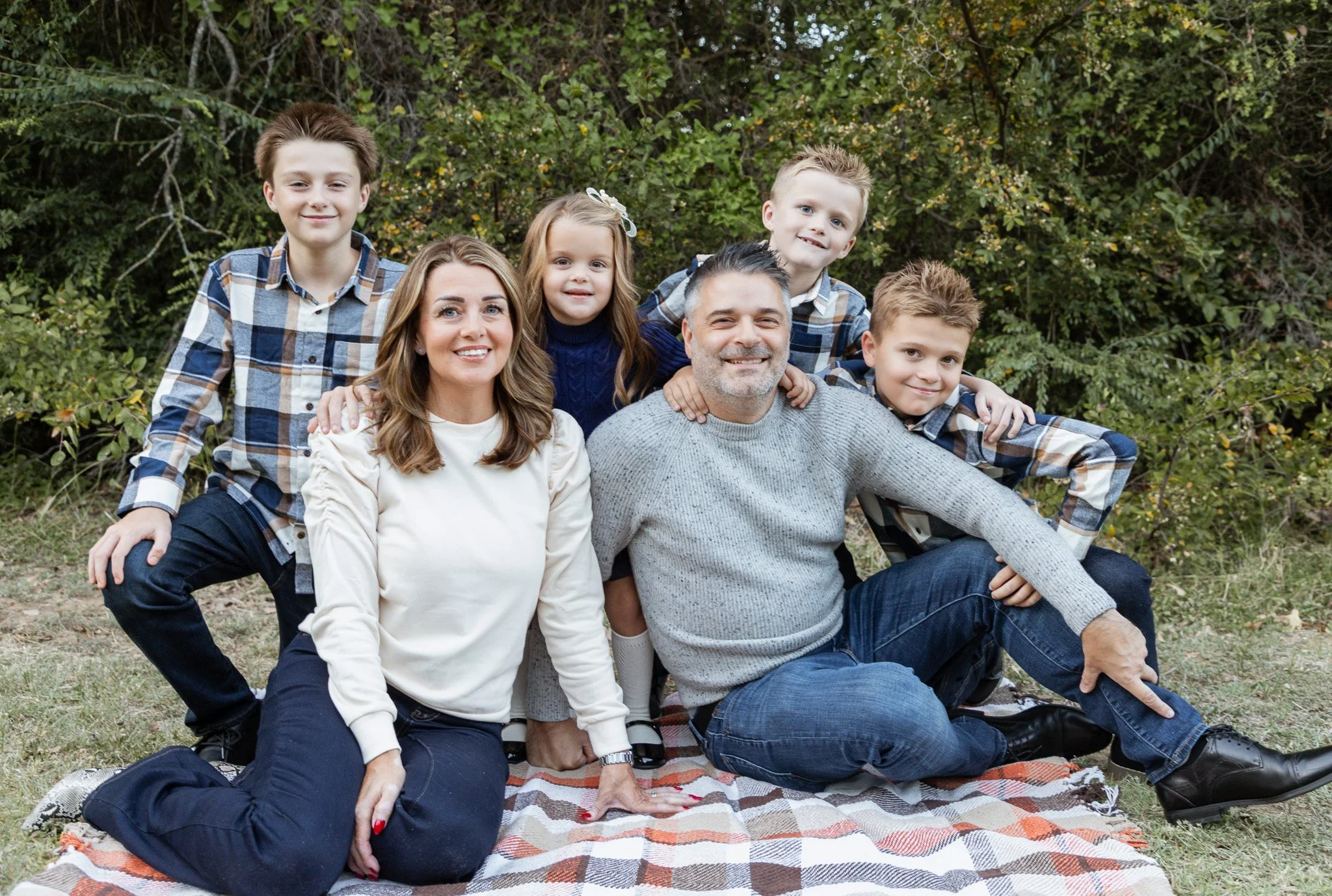 Family of six sitting on a blanket outdoors, smiling with trees in the background.