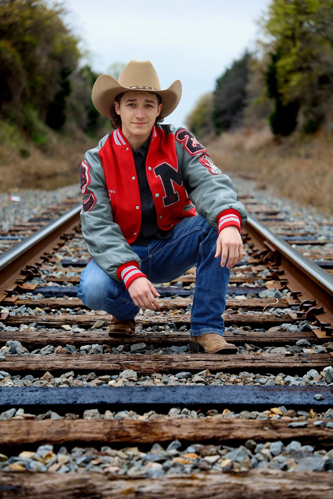 A young man wearing a cowboy hat, red and gray varsity jacket, black shirt, jeans, and brown boots squatting on railroad tracks surrounded by trees and gravel.
