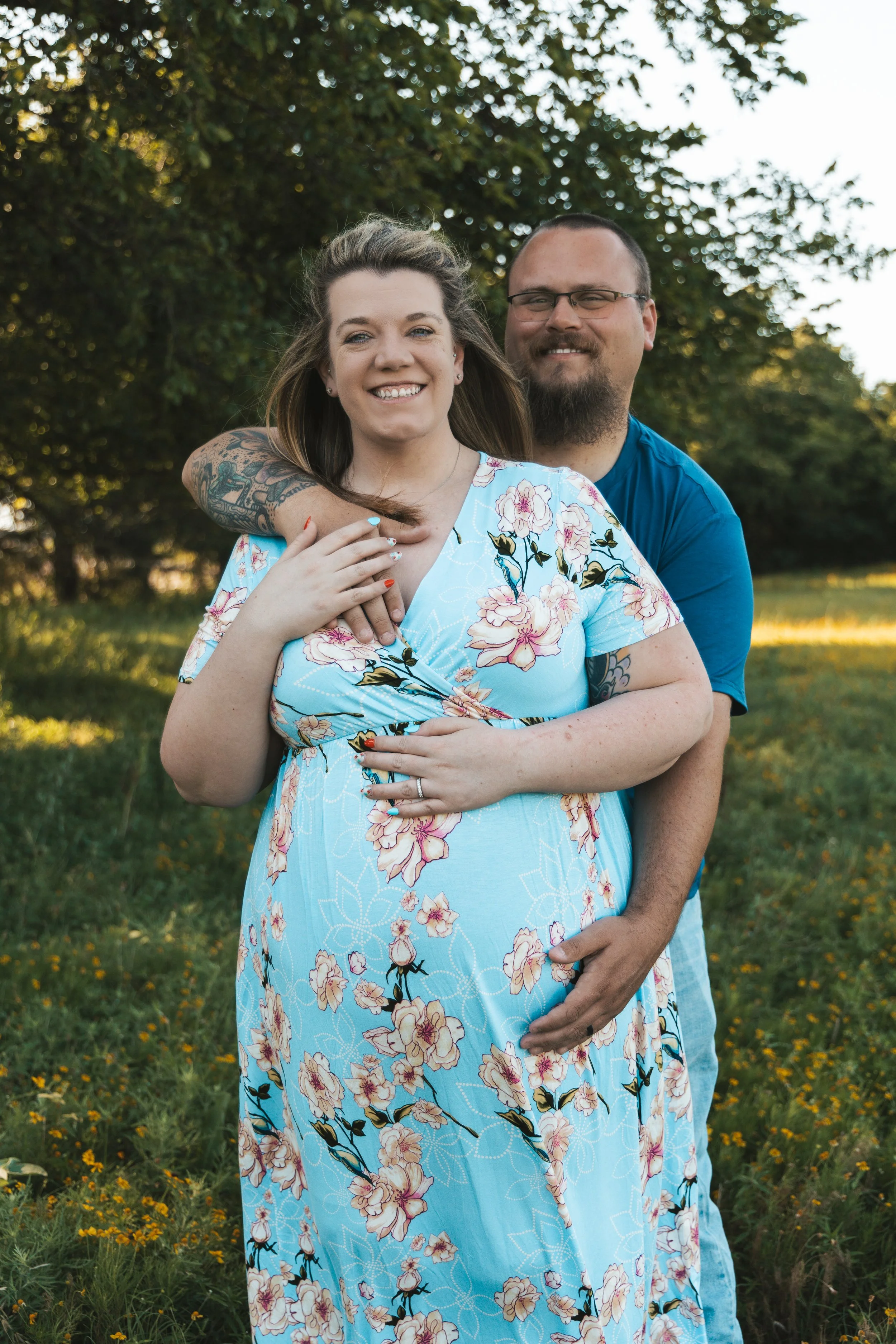 A smiling woman in a blue floral dress stands in front of a man. The man has his arm around her shoulder, also smiling, and is wearing glasses and a blue shirt. They are outdoors in a field with green grass and yellow flowers, with trees in the backg