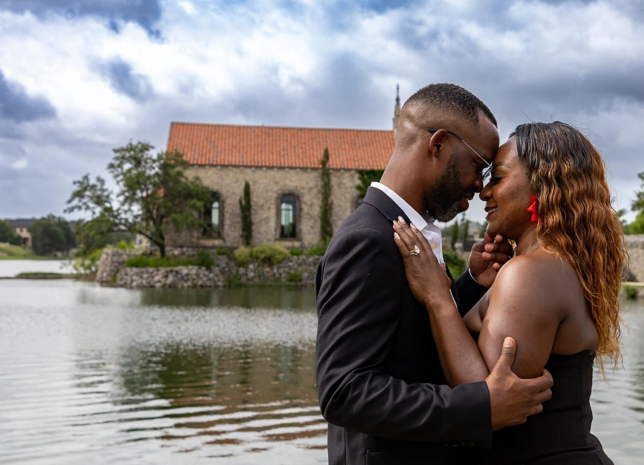 A couple sharing an intimate moment by a river, with a stone building and a cloudy sky in the background.
