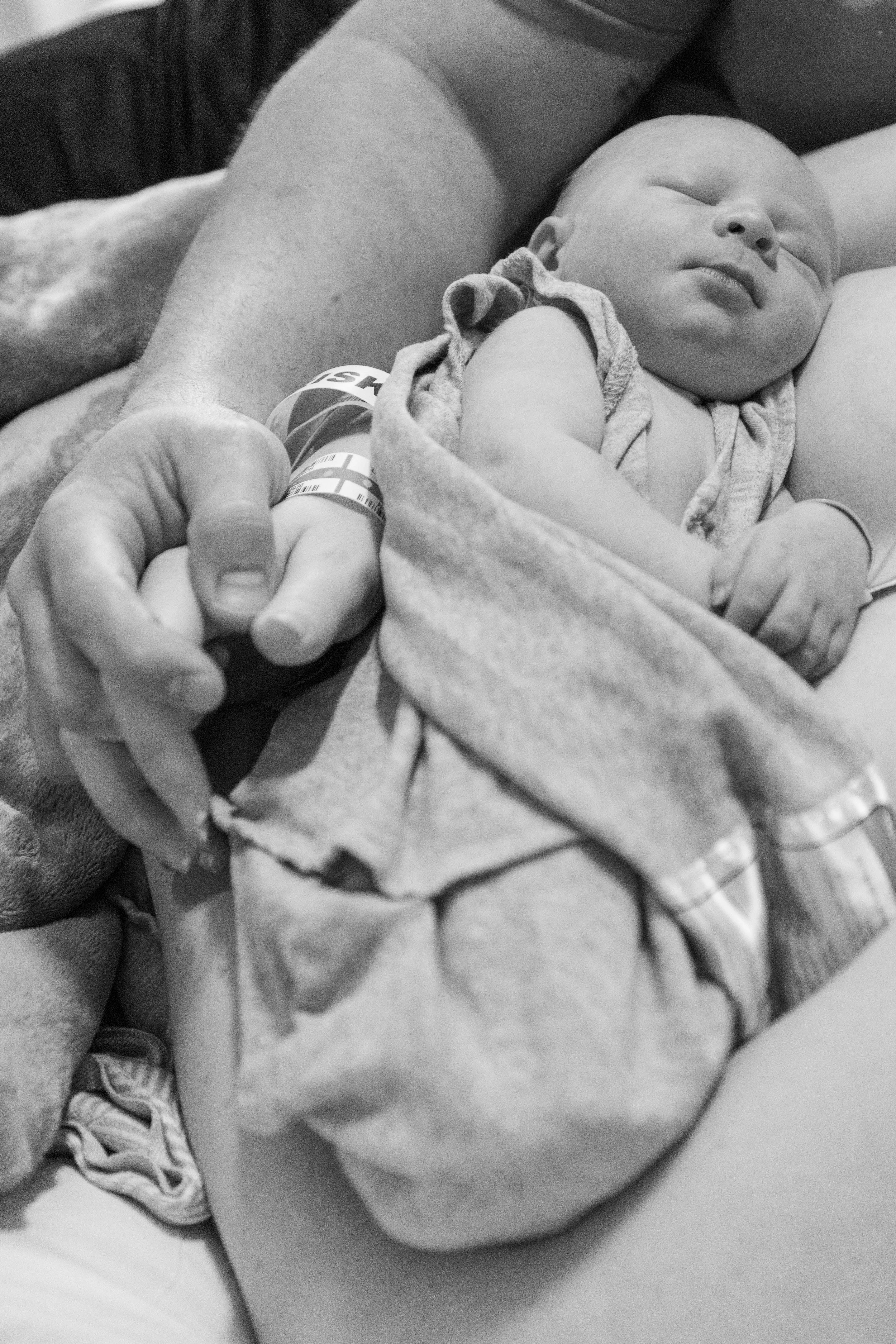 A newborn baby lying on someone's chest, wrapped in a blanket, with an adult's hand gently holding their tiny fingers. The scene suggests a calm, nurturing moment in black and white.