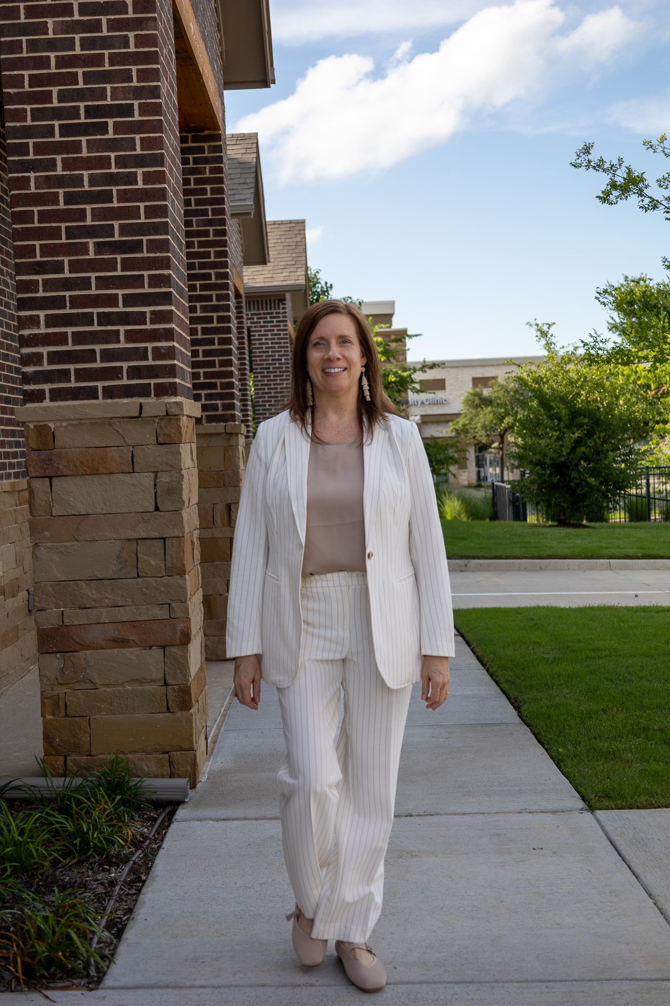 A woman in a white pinstripe suit walking on a sidewalk outside a brick building with green trees and a blue sky in the background.