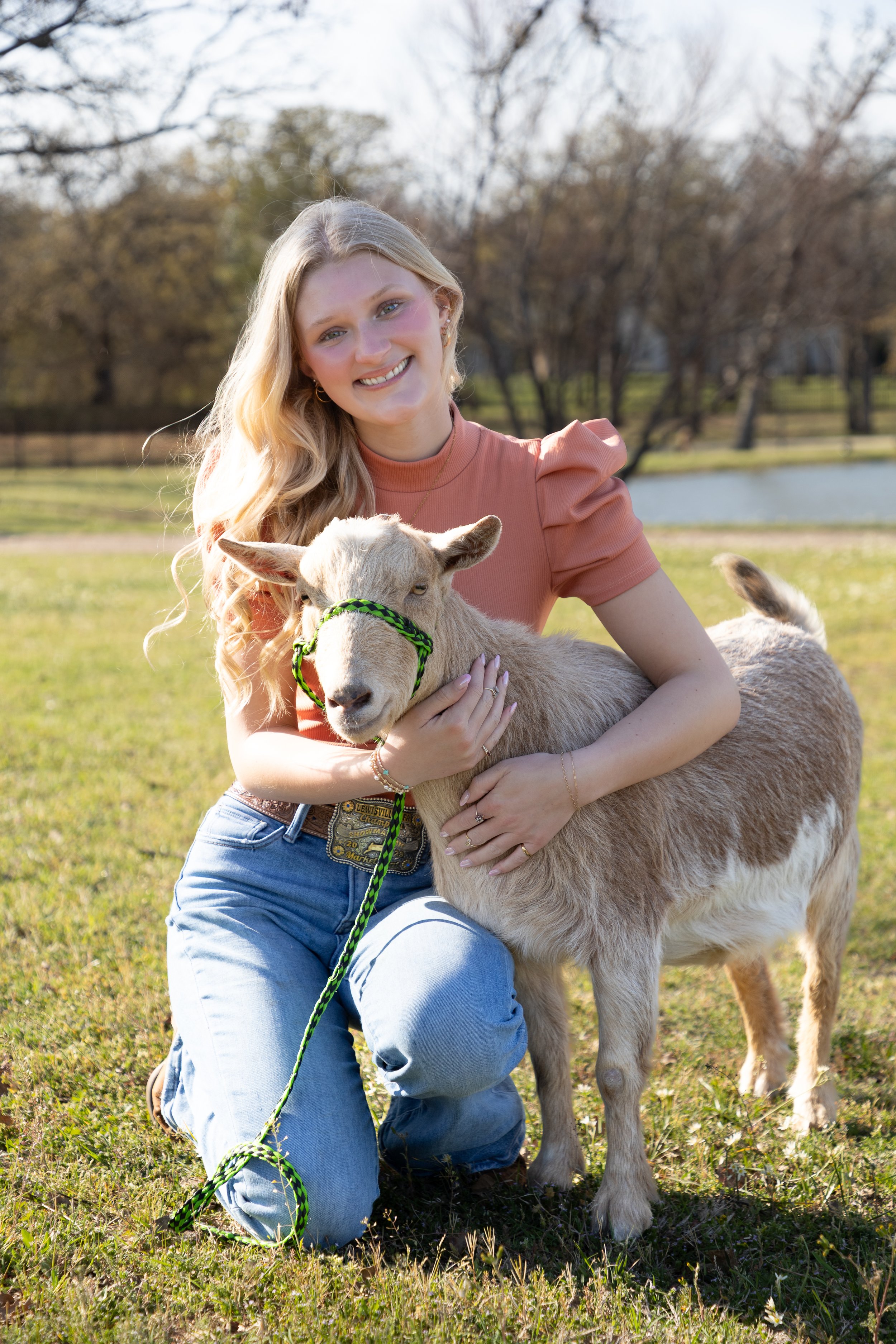 A woman with long blonde hair smiling while hugging a small goat with a green halter, outdoors in a grassy area near a pond, with trees in the background.