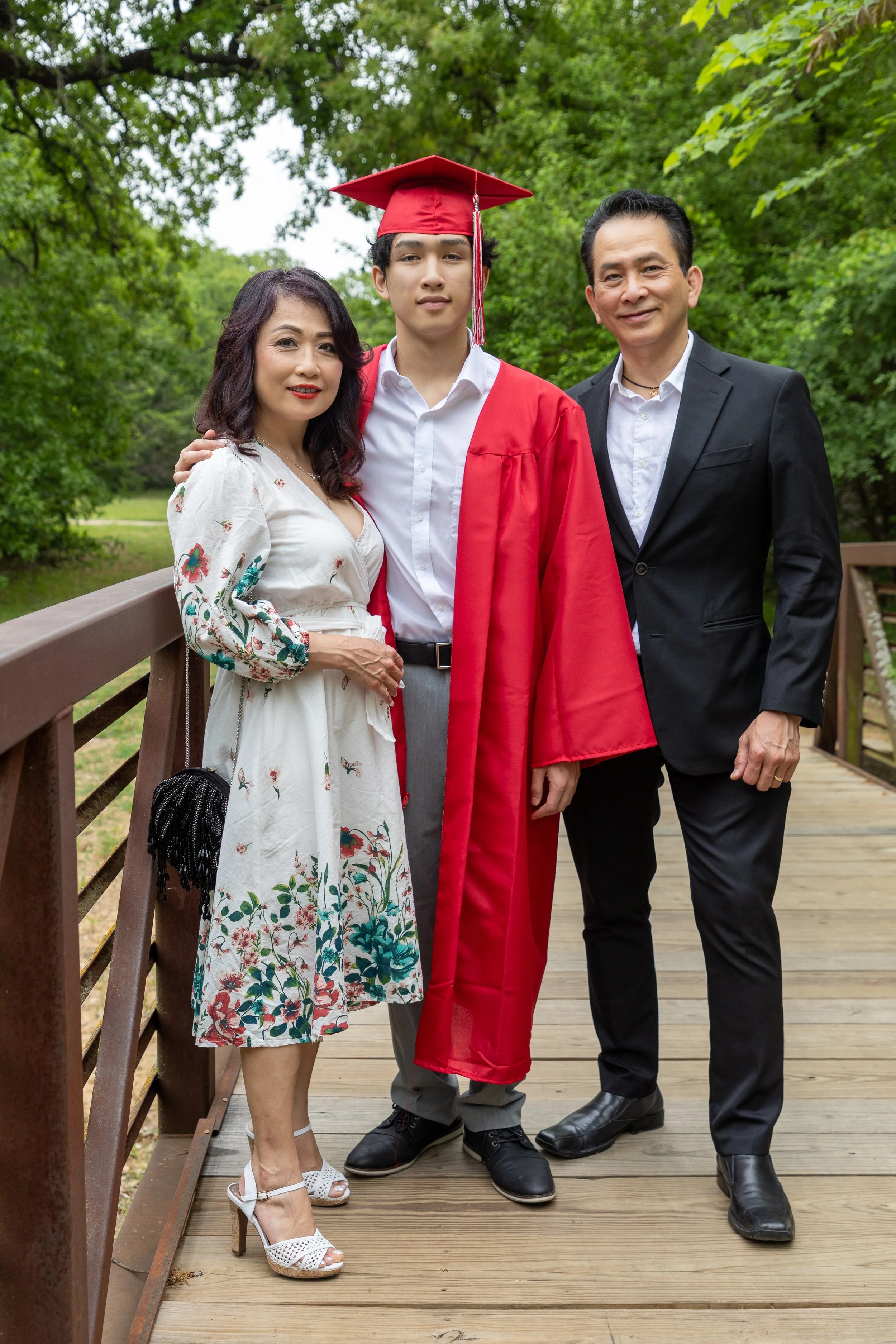 A young man in a graduation cap and gown stands between his mother and father on a wooden bridge outdoors surrounded by green trees.