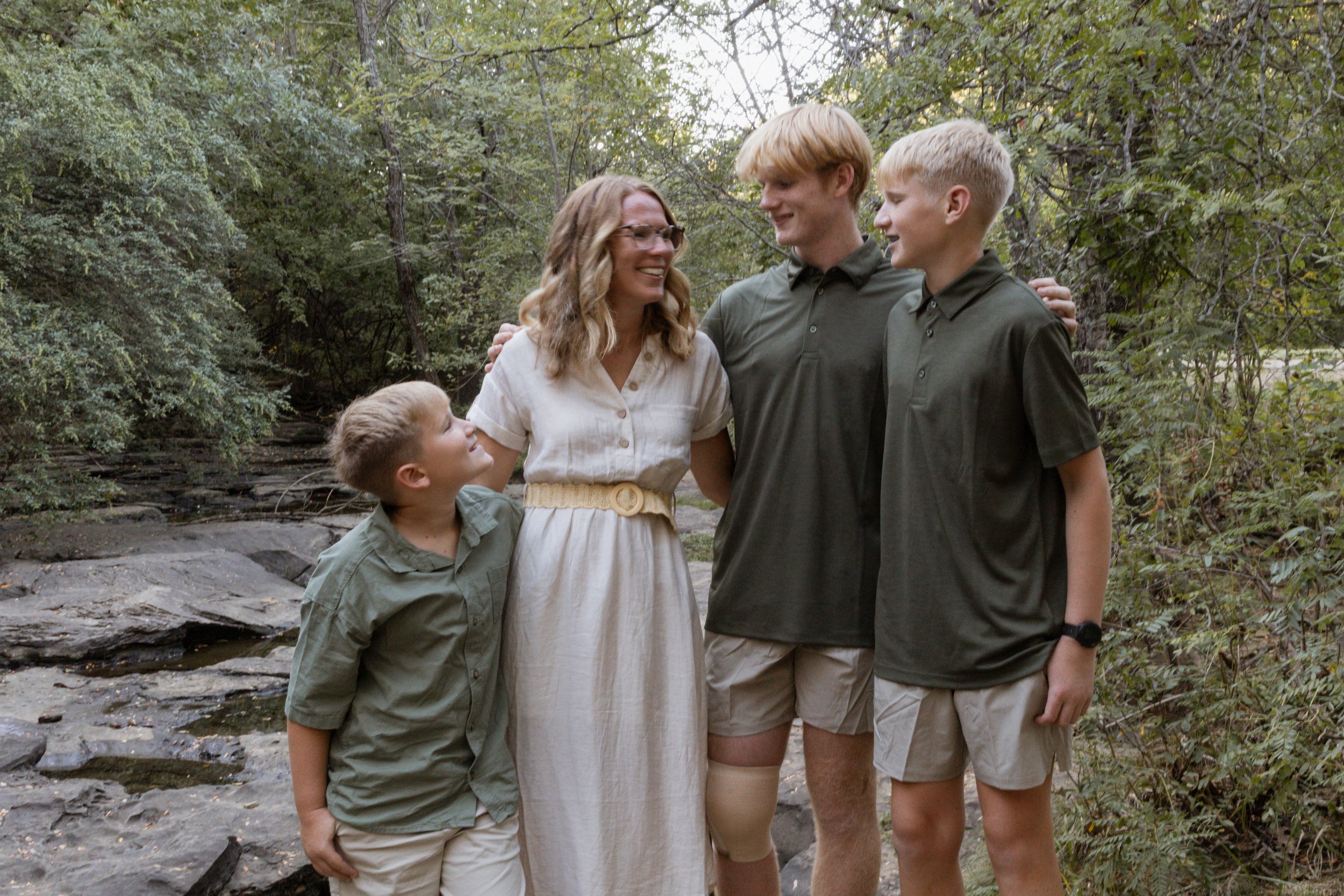 A woman and three boys standing outdoors in a forested area, smiling and looking at each other.