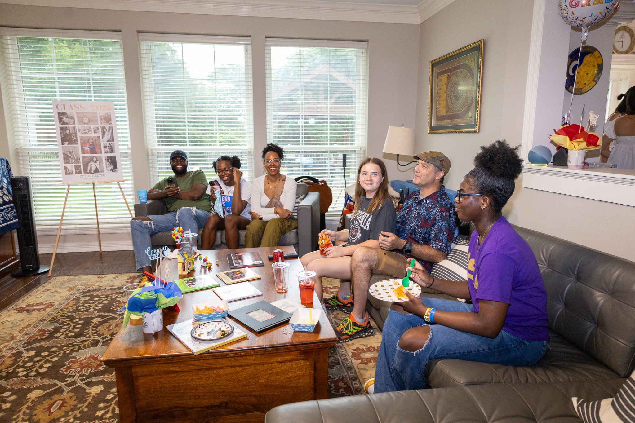 A diverse group of seven people, including children and adults, gathered in a living room for a birthday celebration. They are sitting on couches and enjoying snacks and drinks, with party decorations, balloons, and a 'Congrats' sign visible.