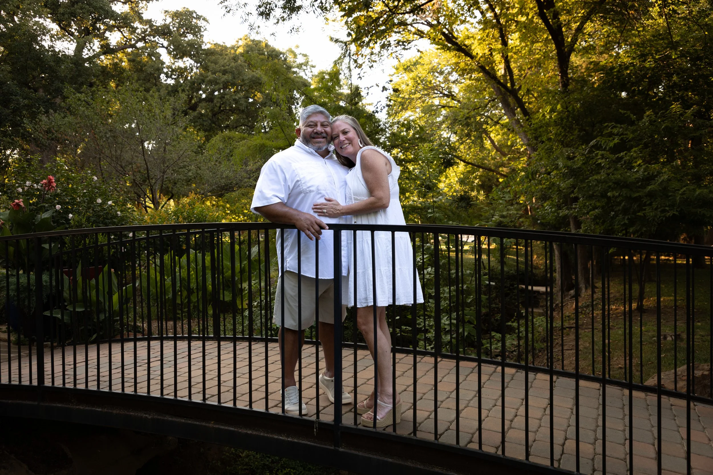A happy couple standing on a small stone bridge in a lush green park, smiling and embracing each other. The man has gray hair and a beard, wearing a white shirt and khaki shorts. The woman has long hair, wearing a white dress and sandals. Sunlight fi