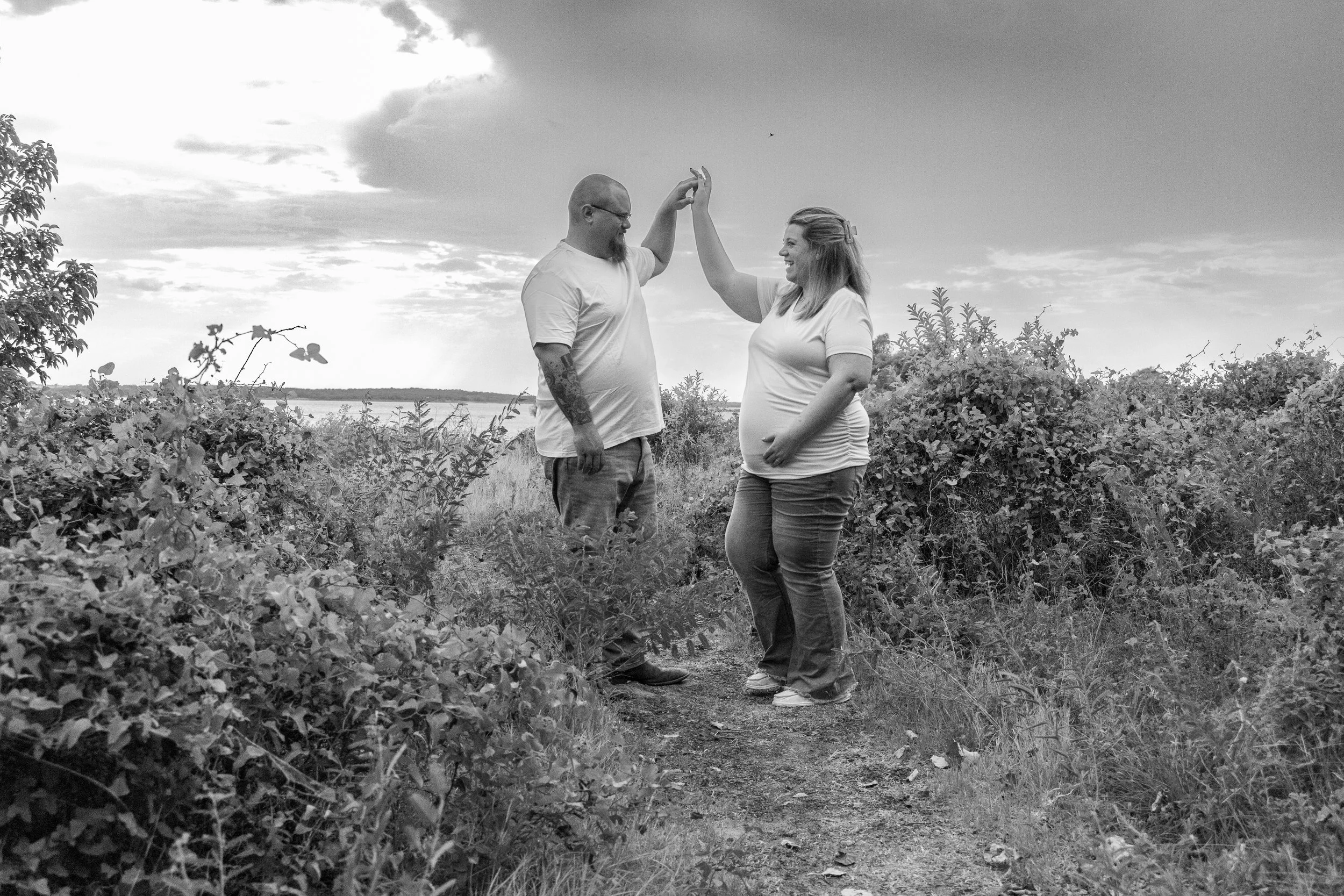 A black and white photo of a man and pregnant woman standing outdoors among bushes, sharing a high-five. The man has a beard, glasses, and tattoos, while the woman has long hair and is smiling. The sky is partly cloudy.