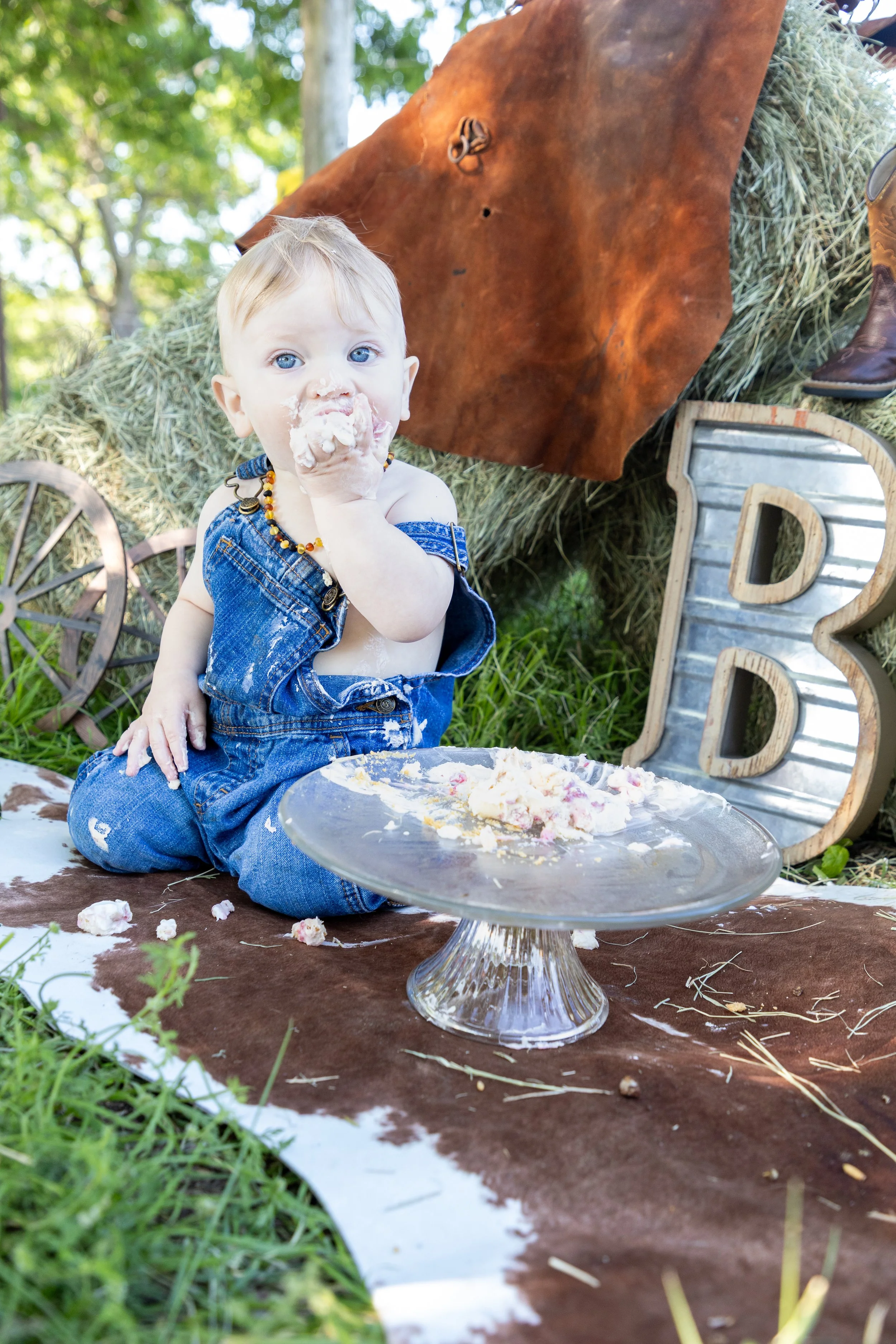 A young child with blonde hair and blue eyes, wearing denim overalls, sitting on a brown and white cowhide rug outside, eating cake with white frosting and red cherries. The child is covered in frosting. In the background are hay bales with a leather