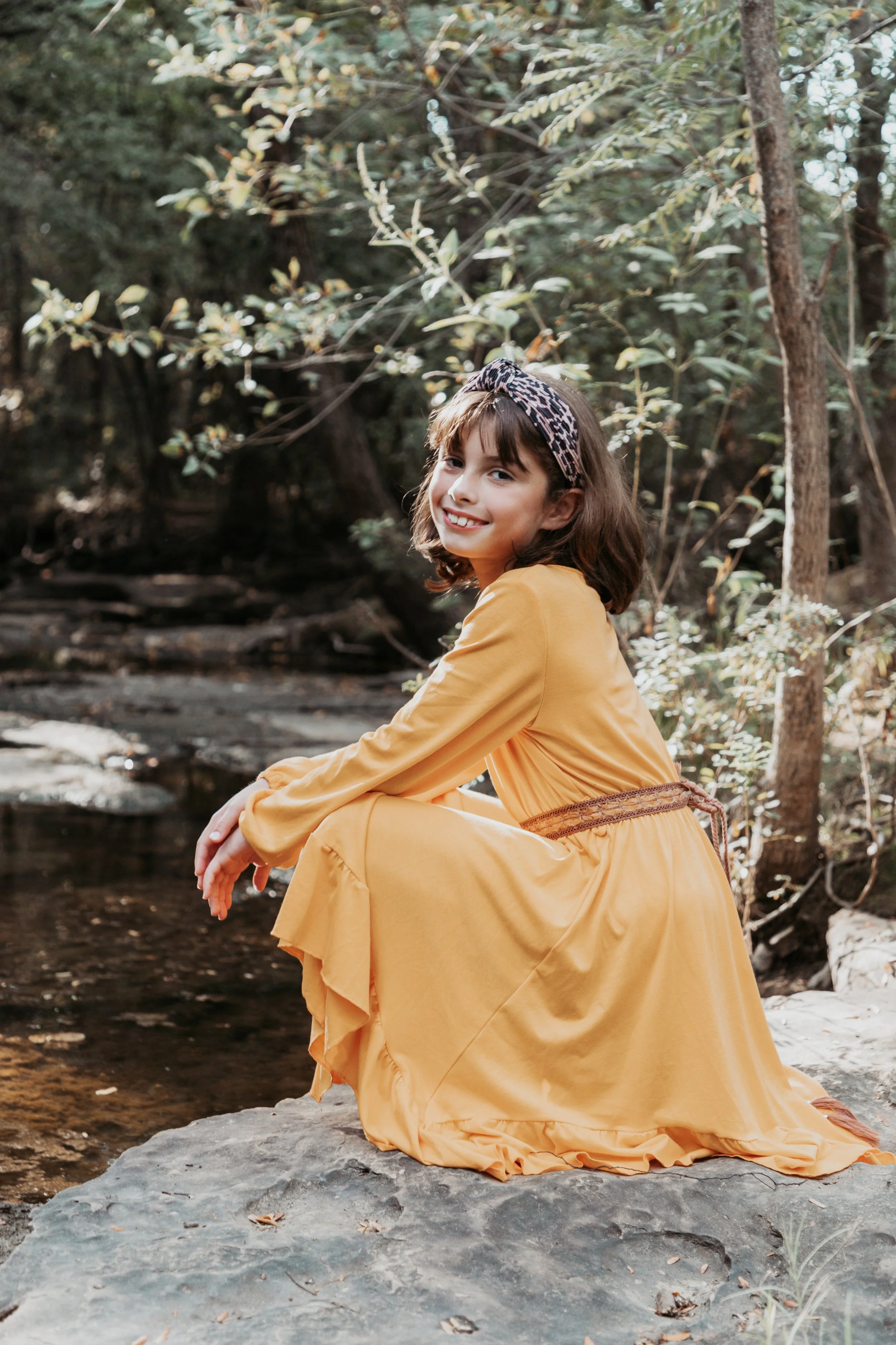 A young girl sitting on a rock by a stream in a forest, smiling at the camera. She wears a long yellow dress and a leopard print headband.