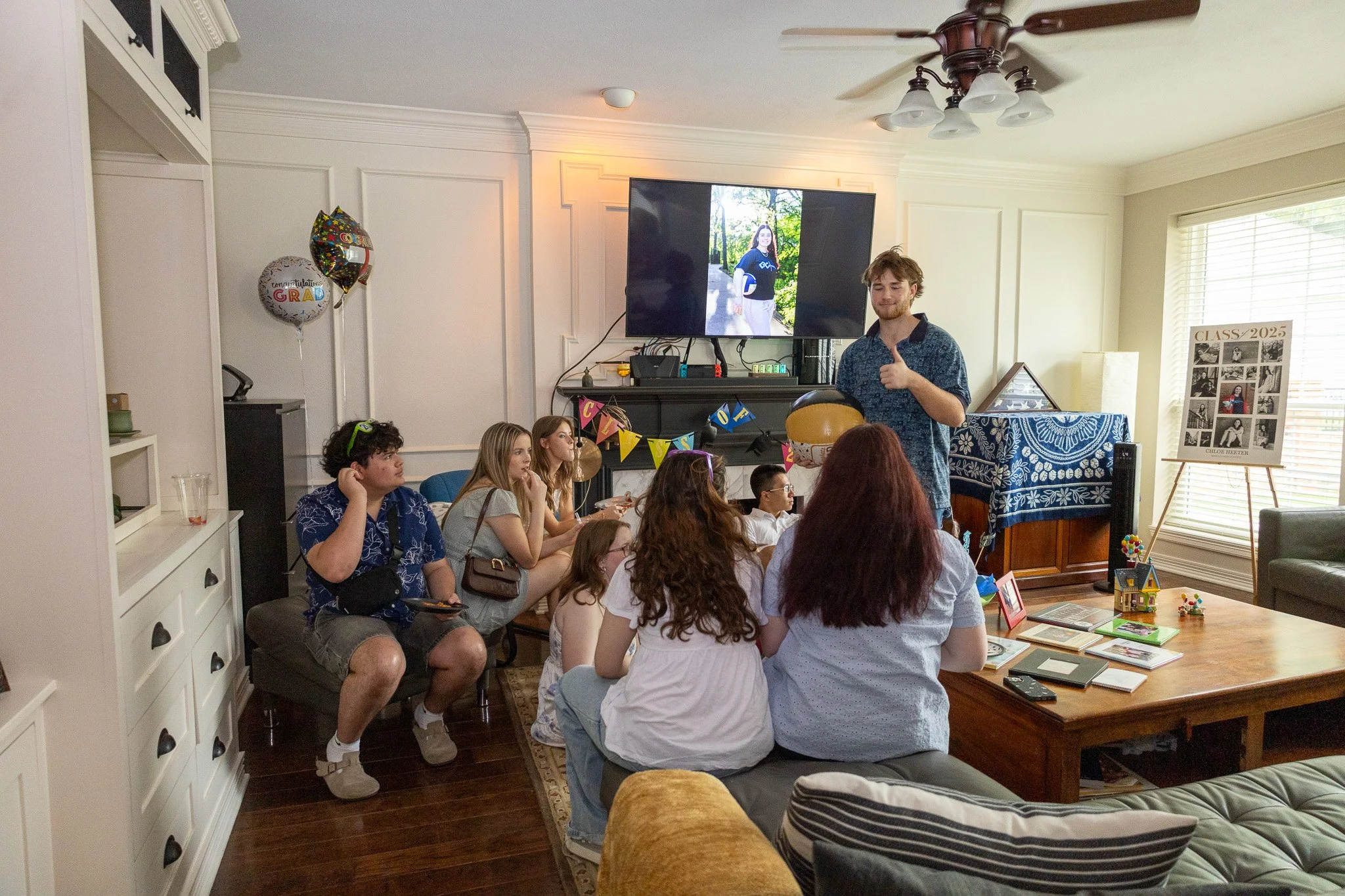 A group of young people gathered in a living room for a graduation celebration. Some are sitting on a bench and others on a couch, watching a man speaking and giving a thumbs up. The room is decorated with balloons, a banner, and a collage marked 'Cl
