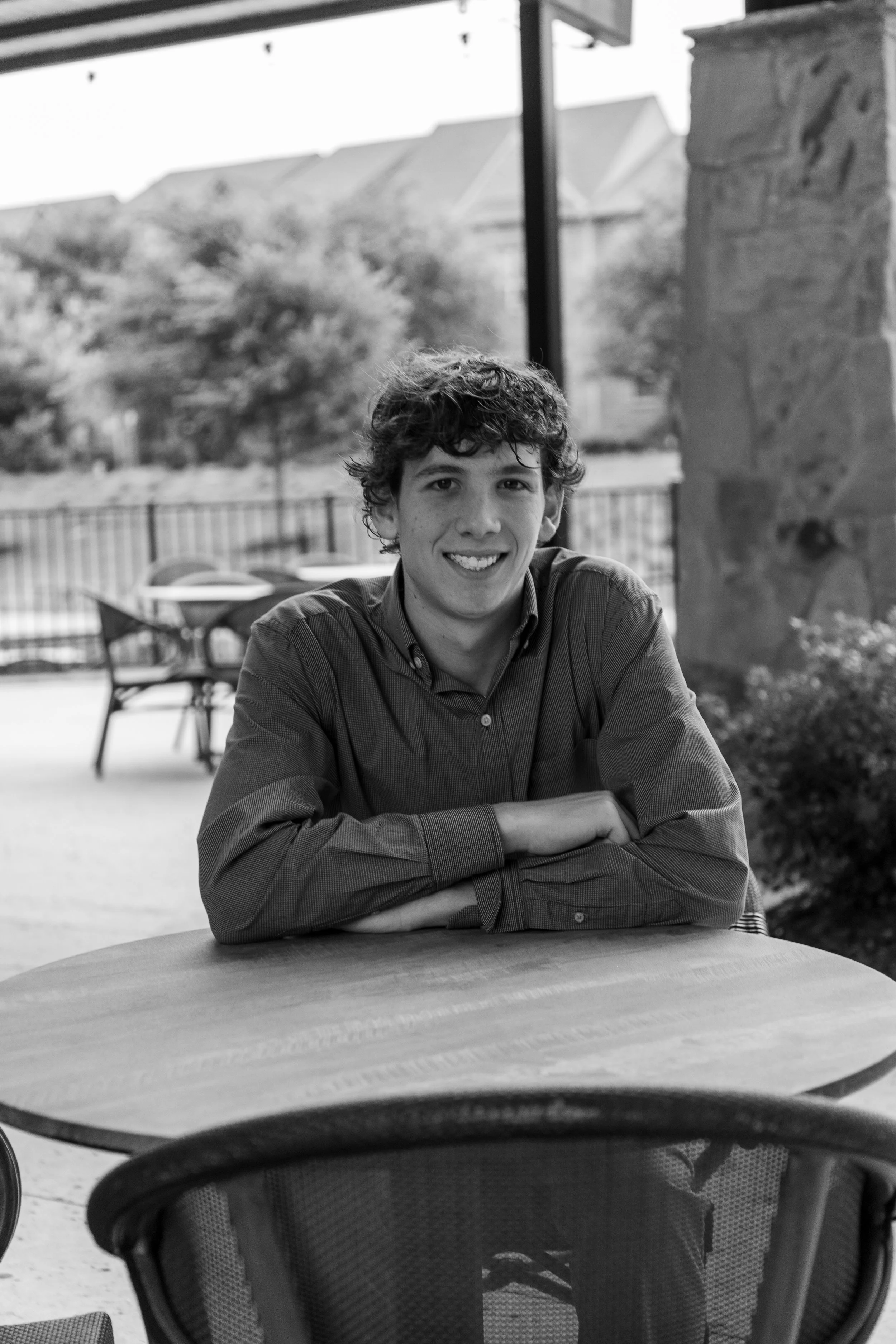 A young man with curly hair smiling, sitting at a round table outdoors, with chairs and trees visible in the background.