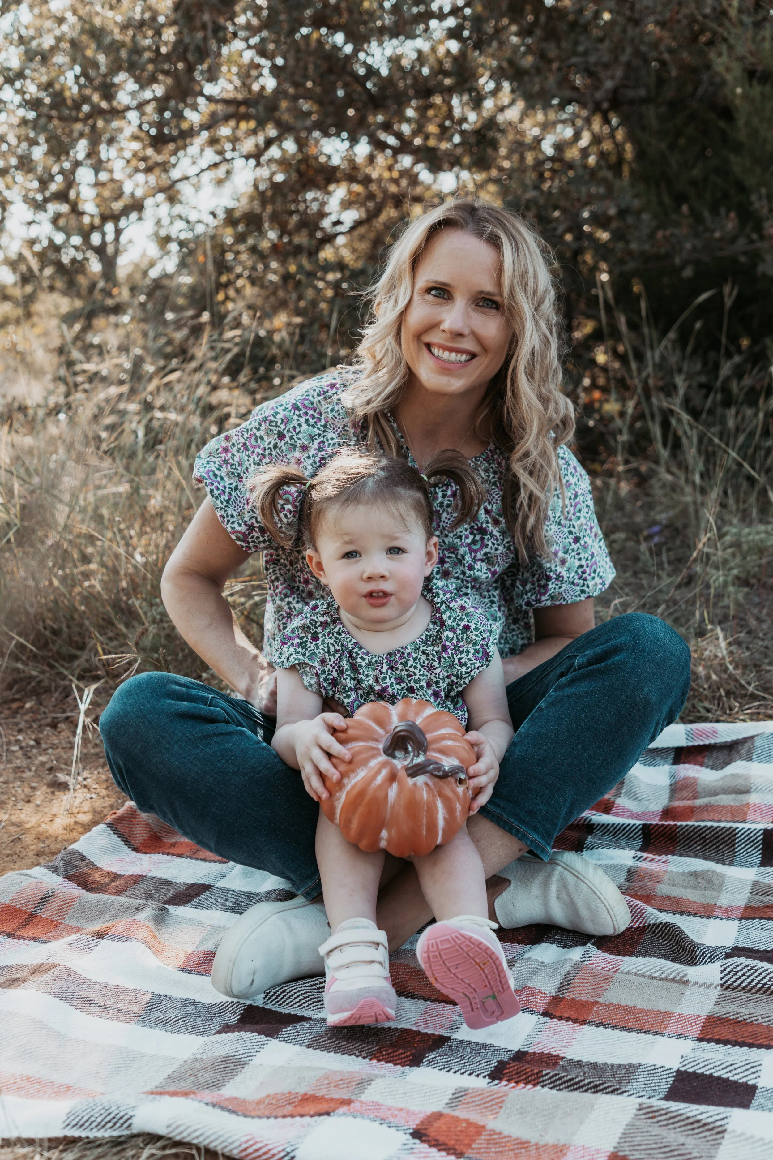 A woman and a young girl sitting on a plaid picnic blanket outdoors, holding a small pumpkin, surrounded by trees and grass, during daytime.