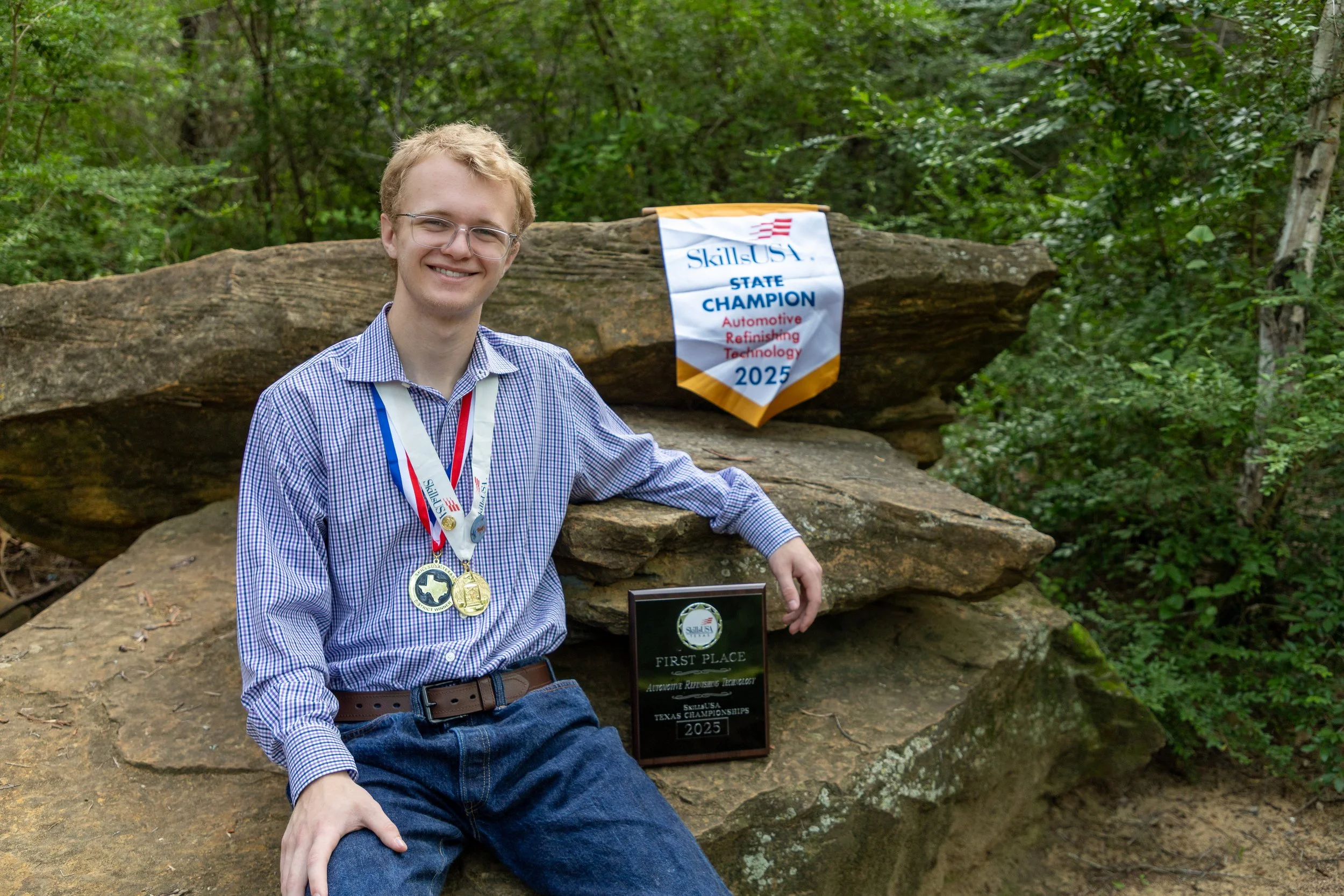 Young man smiling, wearing glasses, a checkered shirt, and medals around his neck, sitting on a large rock in a wooded area, with a first-place plaque and a SkillsUSA banner behind him that reads 'State Champion 2025' in Automotive Refinishing Techno