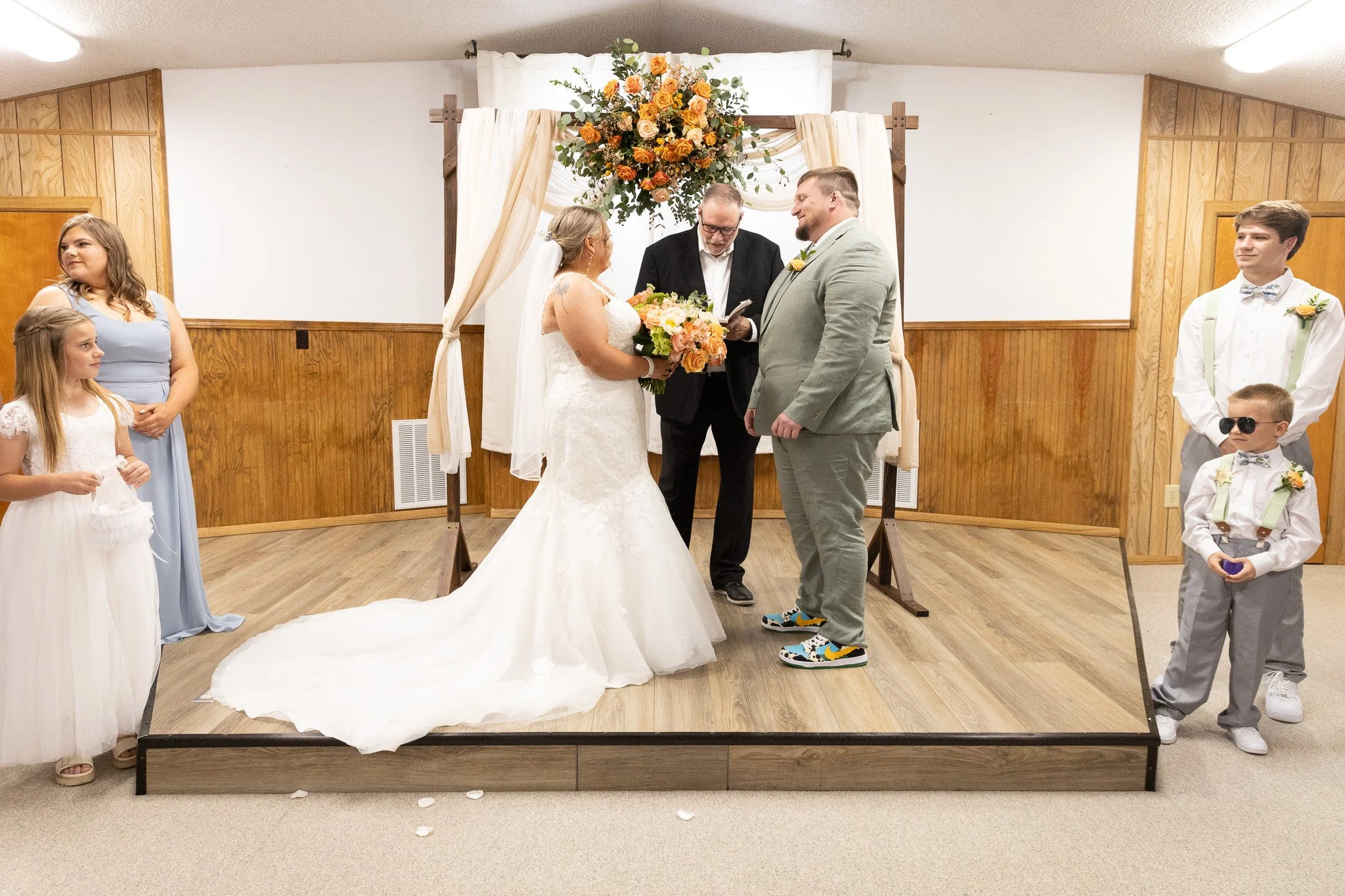 Wedding ceremony with a bride and groom standing under a floral arch, exchanging vows. The bride is wearing a white wedding gown and holding a bouquet, and the groom is in a gray suit with colorful sneakers. An officiant is reading from a book. On th