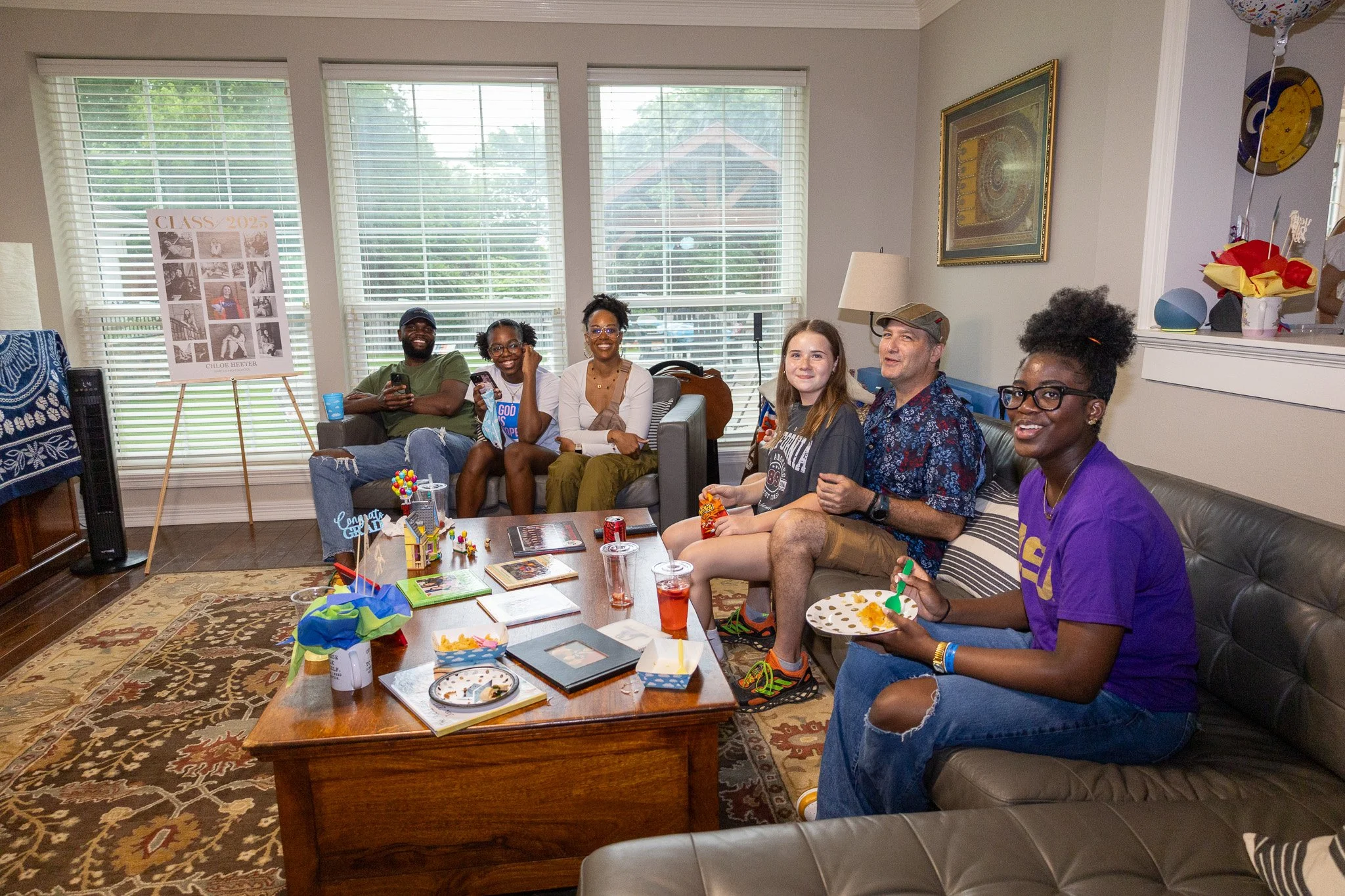 A diverse group of seven people sitting on a couch and chairs, enjoying a gathering in a living room decorated with family photos, artwork, and decorations.