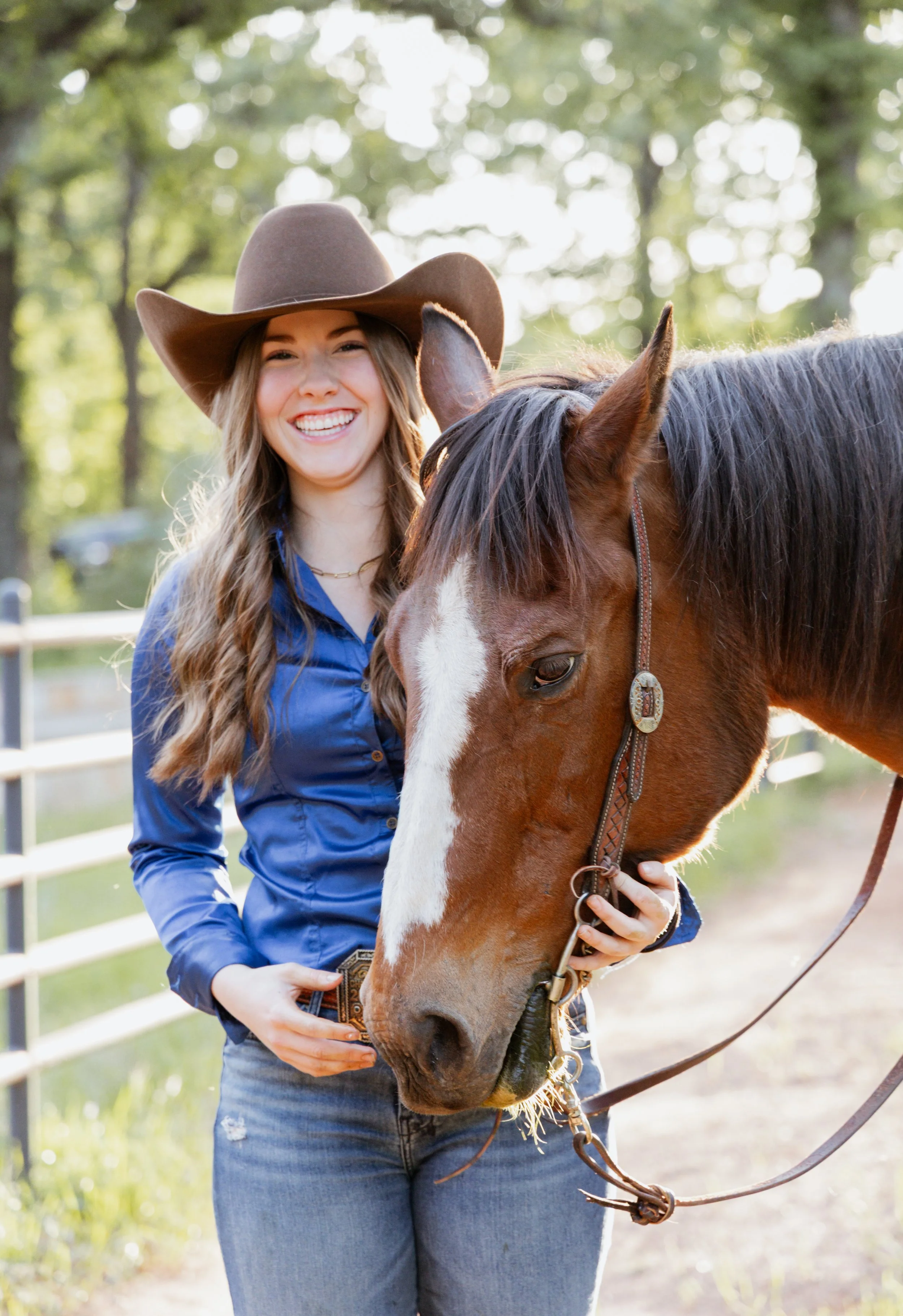 A woman wearing a cowboy hat and blue shirt smiling as she stands next to a brown horse with a white face marking outdoors in a green, wooded area.