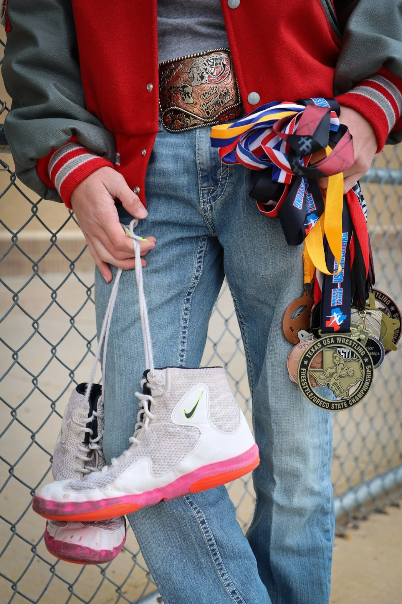 Person holding multiple wrestling medals and a pair of Nike high-top sneakers hanging from their hand. The person is wearing a red and green varsity jacket, blue jeans, and a championship belt around their waist, standing next to a chain-link fence.