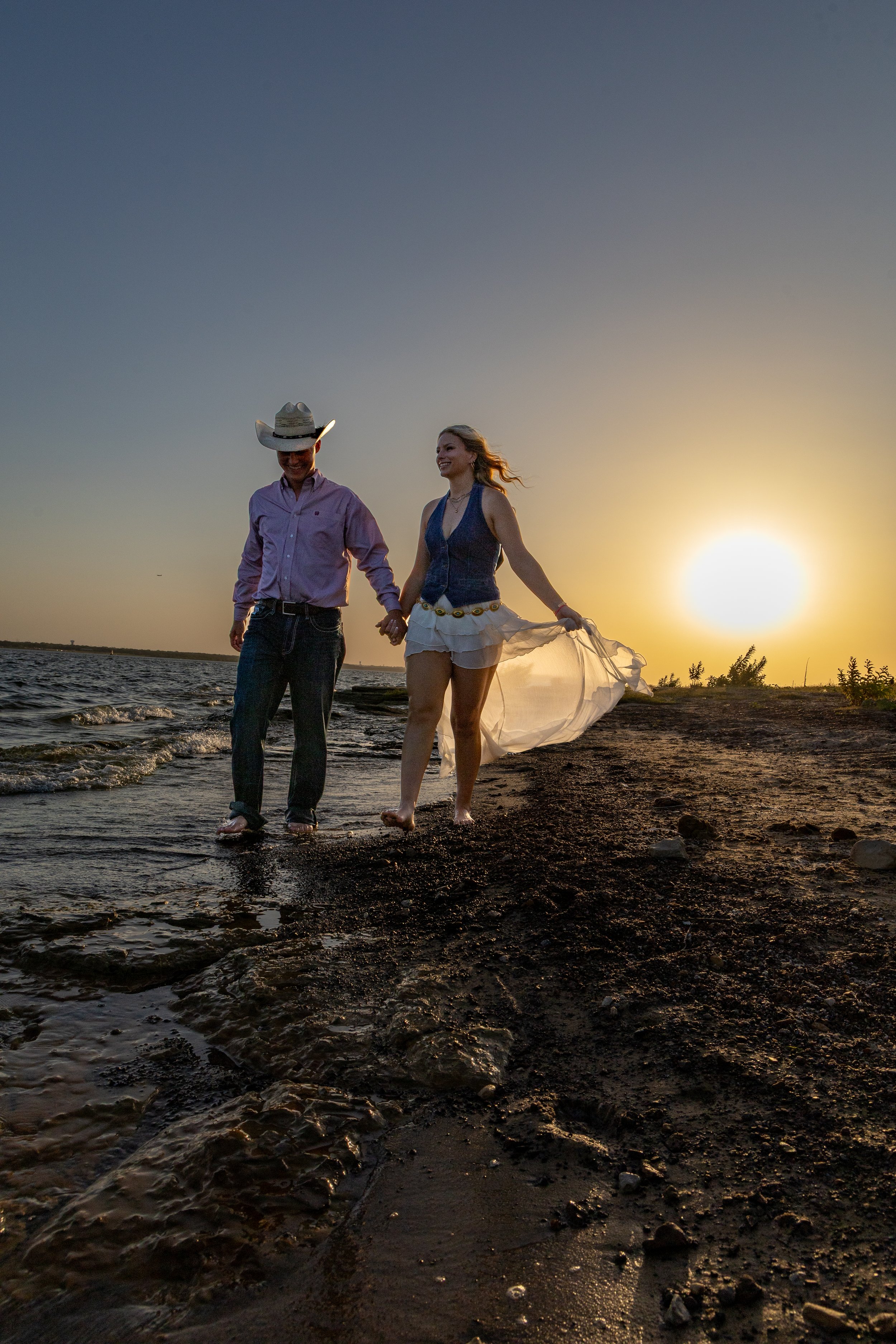 A couple walking hand in hand along the shoreline during sunset, with water gently lapping at their feet and the sky glowing with warm colors.