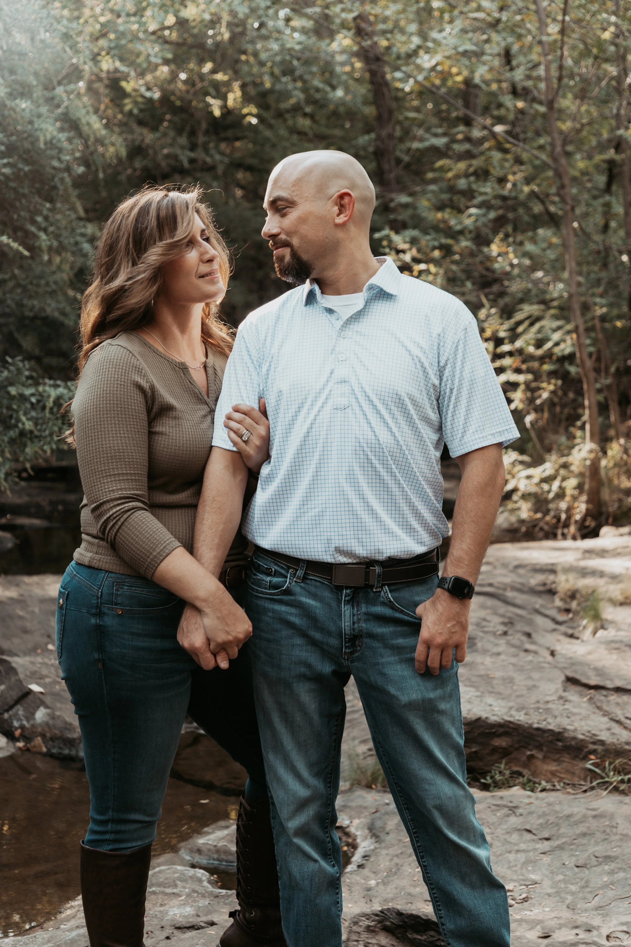 A couple standing closely outdoors near a stream, holding hands and gazing at each other, surrounded by trees with green leaves.