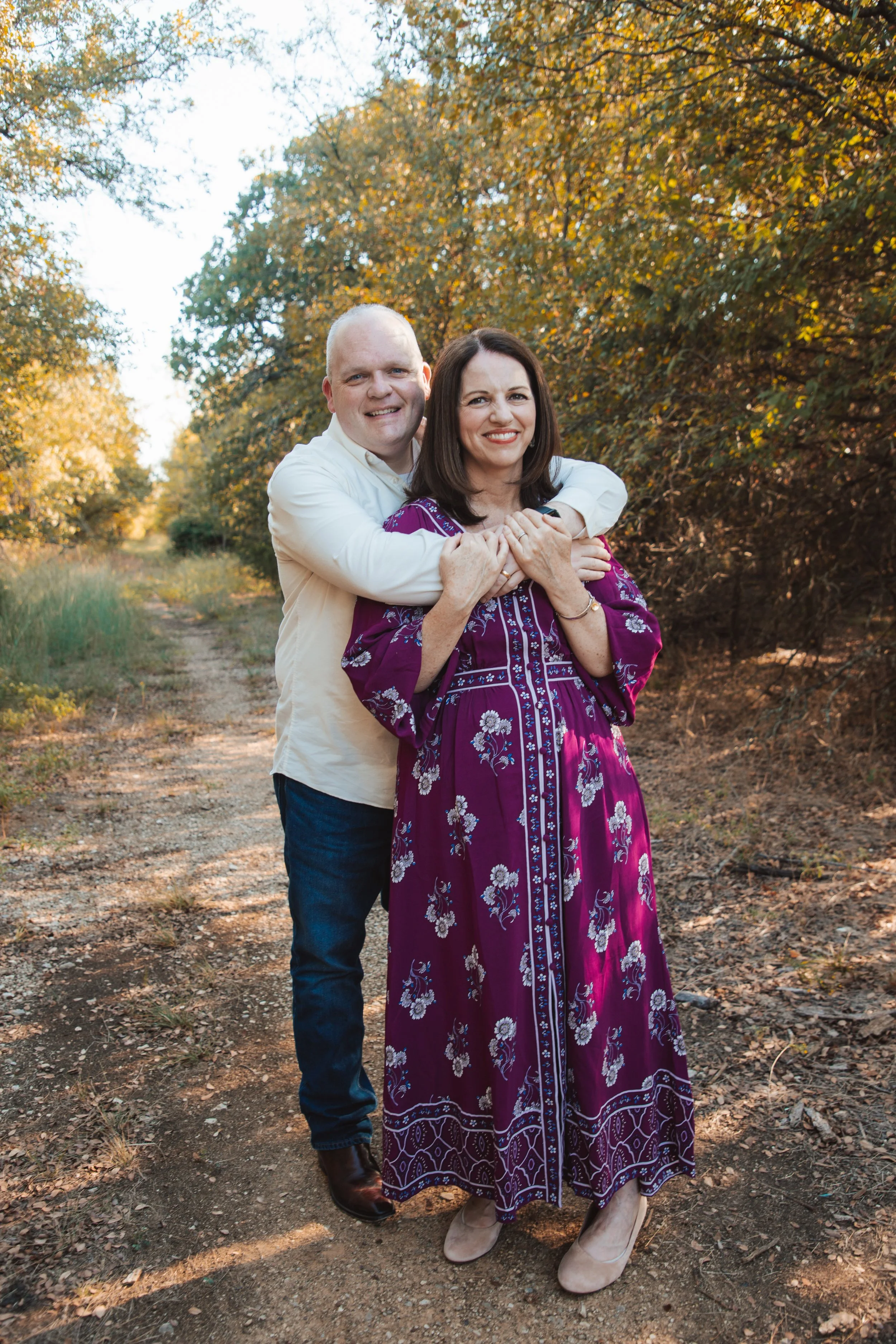 A man and a woman standing outdoors on a dirt path surrounded by trees with autumn foliage. The man is hugging the woman from behind, and they are both smiling at the camera. The woman is wearing a long purple floral dress, and the man is dressed in 