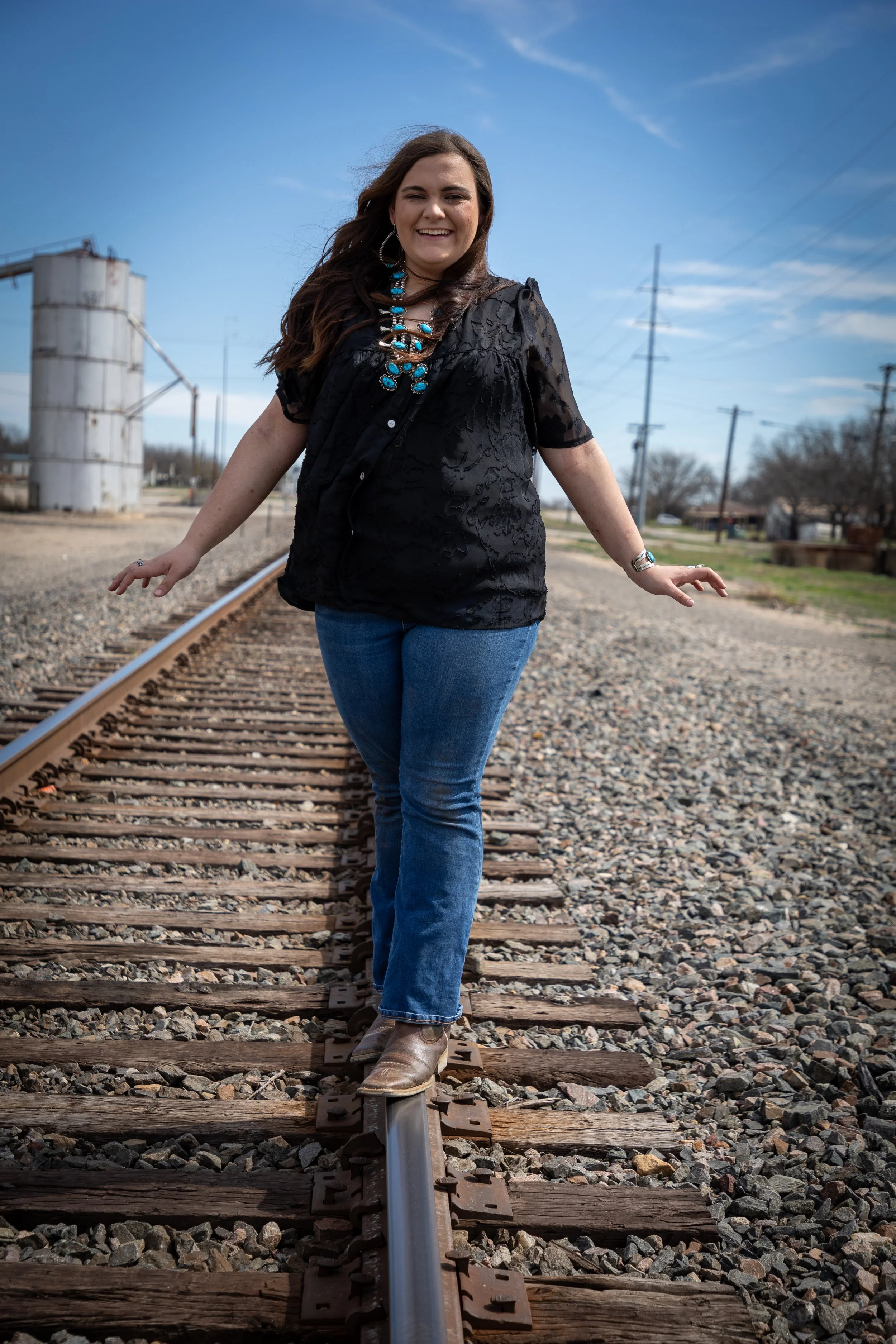 A woman walking on railway tracks outdoors on a sunny day, wearing a black top, blue jeans, and boots, smiling at the camera.