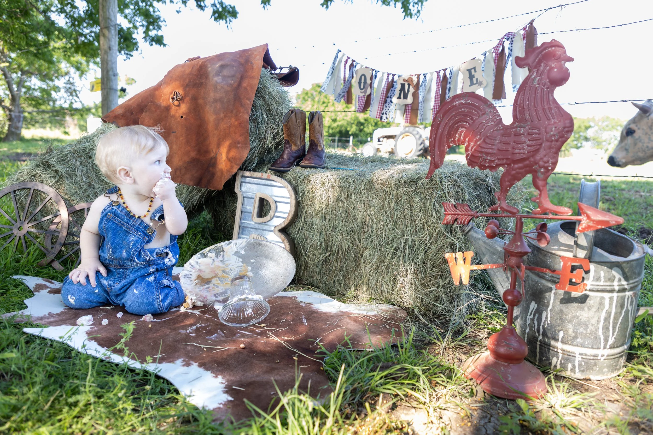 A young child dressed in overalls sitting on the ground outdoors with a pie dish and spoon, surrounded by rustic farm-themed decorations including hay, cowboy boots, a metal chicken weather vane, antique wagon wheels, a miniature wagon, and a banner 