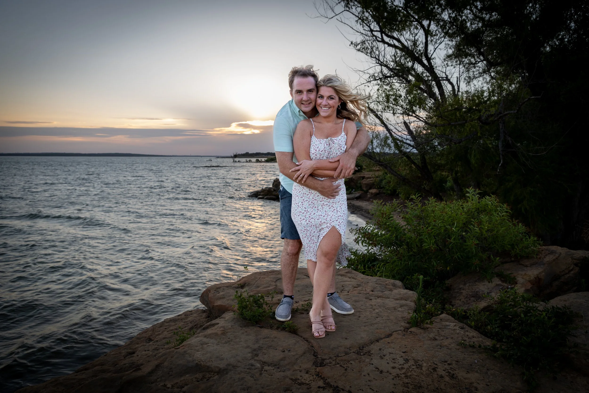 A smiling couple hugging on a rocky shoreline at sunset, with water, trees, and sky in the background.