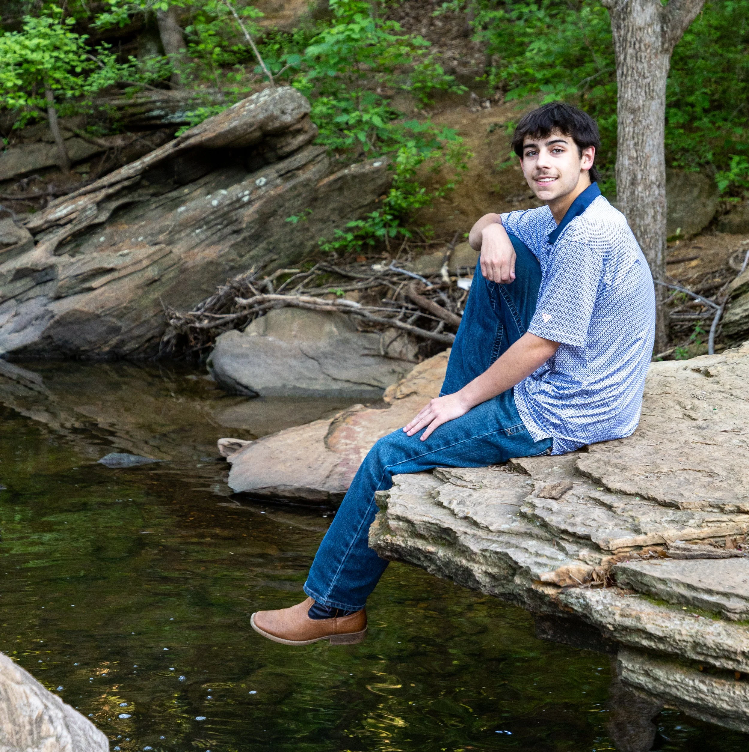A young man with dark hair, wearing a light blue short-sleeved button-up shirt, blue jeans, and tan shoes, sits on a large rock by a creek, surrounded by green trees and foliage.