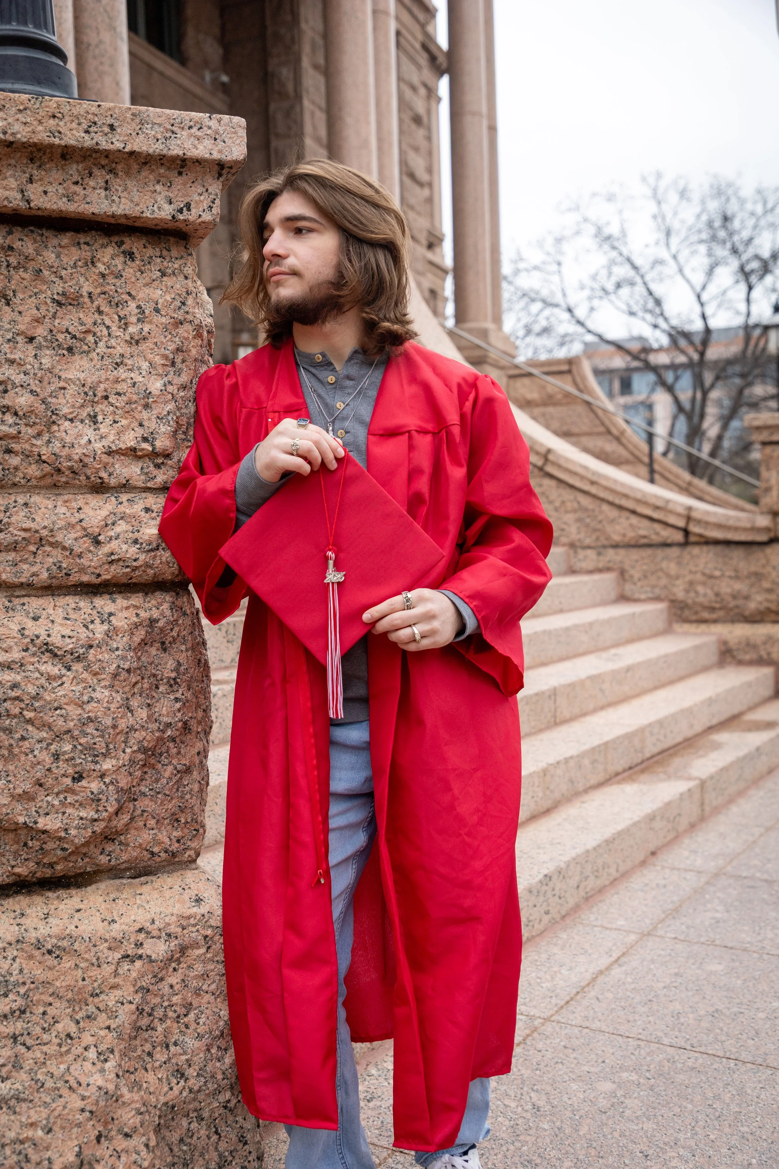 Young man in gray shirt and red graduation gown holding a matching cap outside a historic building with stone stairs and columns.