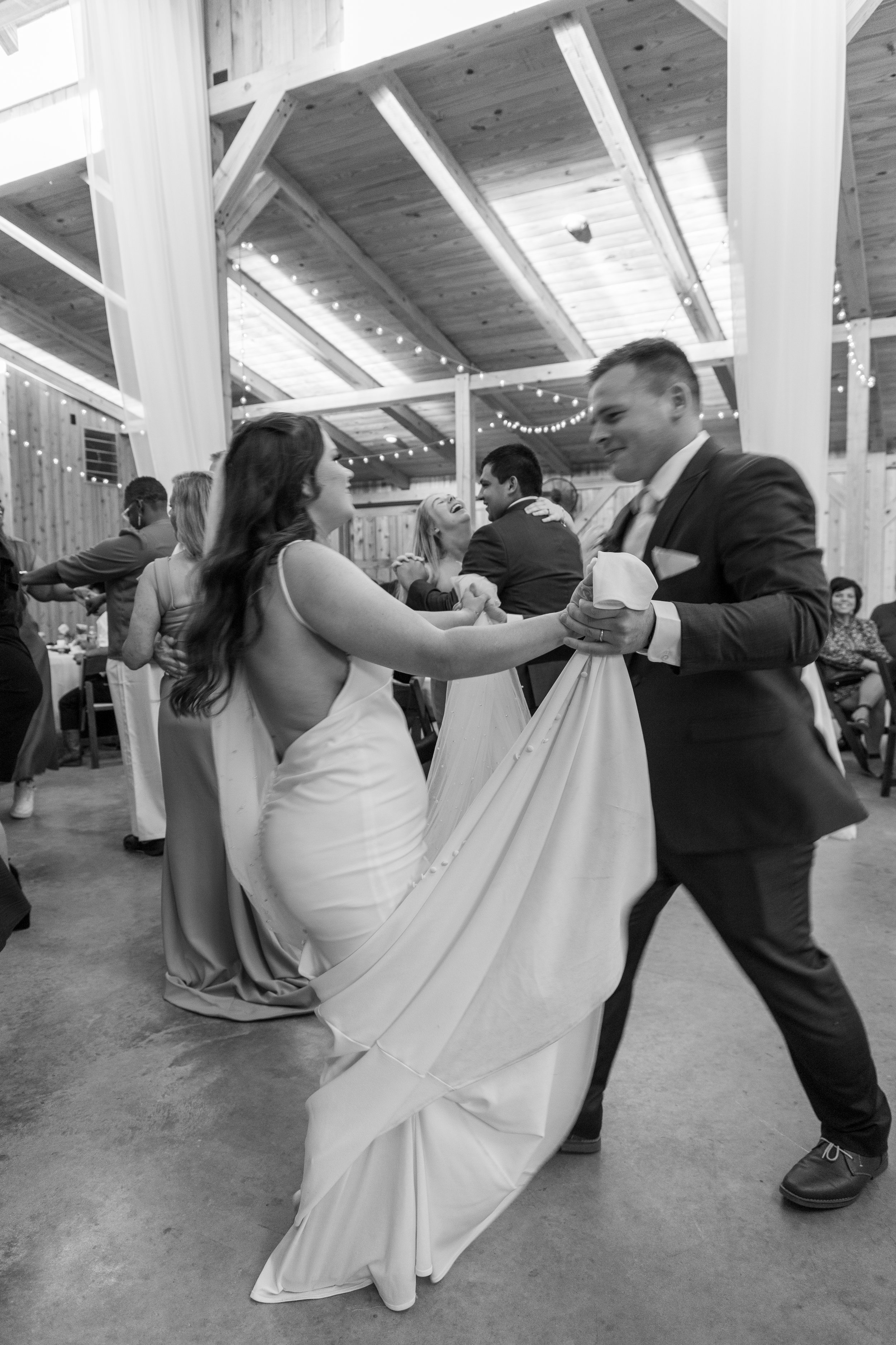 Black and white photo of a wedding reception with a bride and groom dancing together. The bride is holding the bottom of her wedding dress while dancing with the groom. Other guests are dancing and sitting in the background inside a wooden venue with