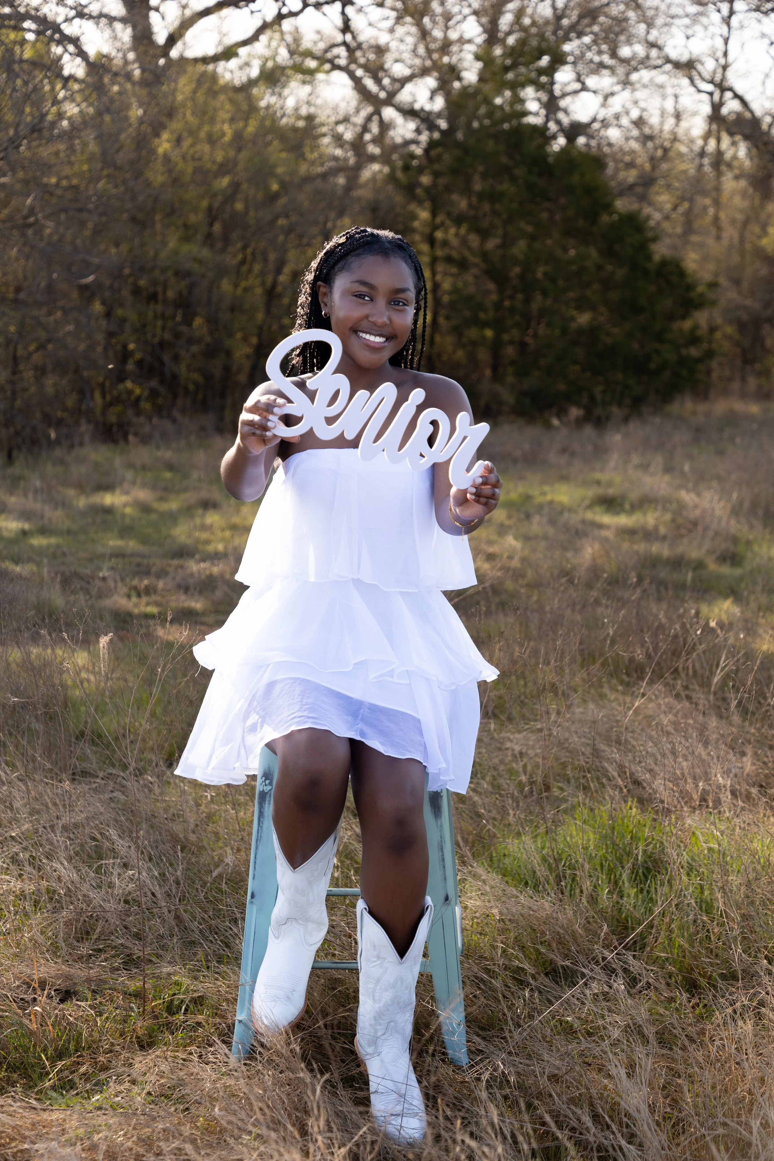 A woman in a white dress and cowboy boots sitting on a light blue ladder outdoors, holding a sign that reads 'Senior', with trees and grassy field in the background.
