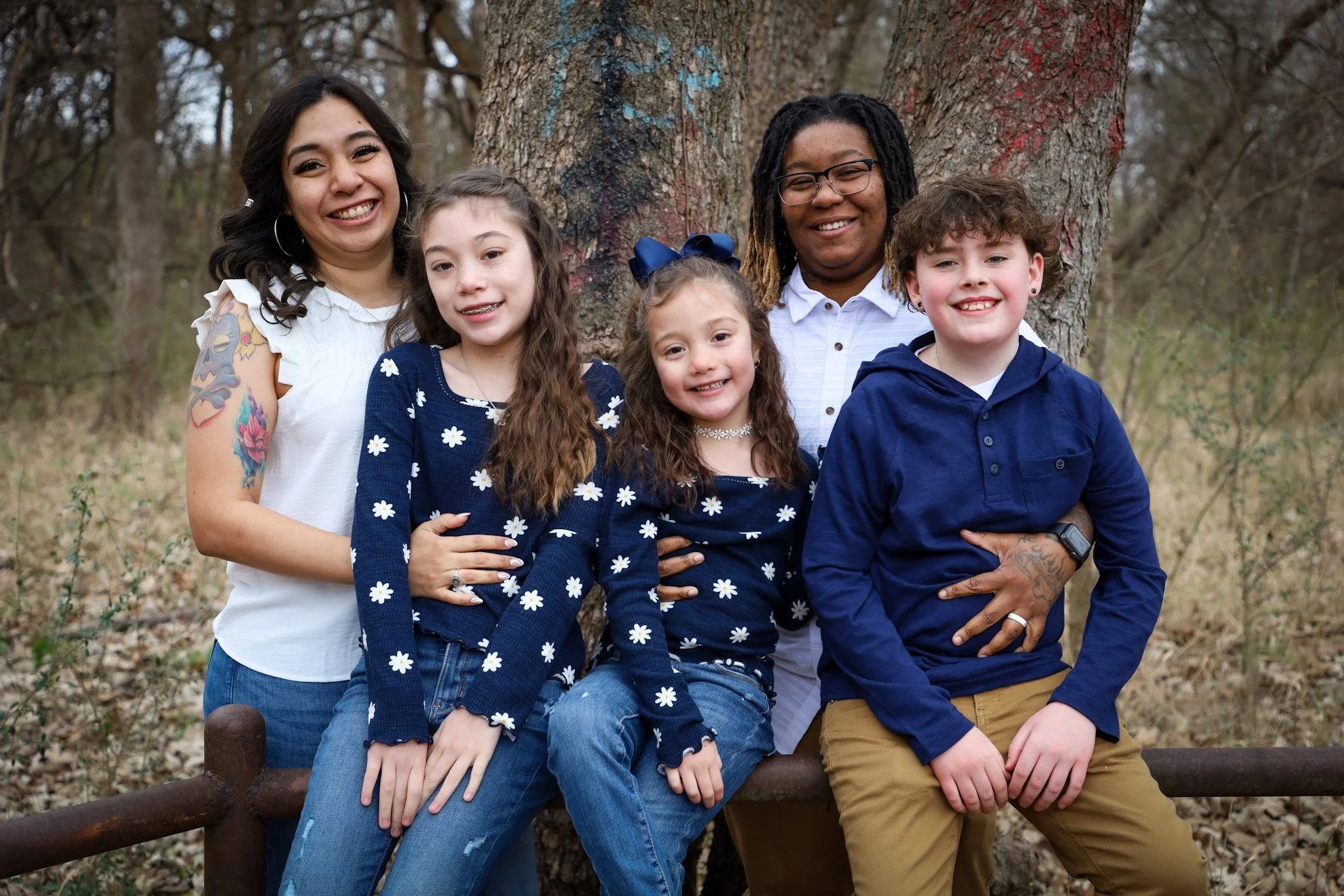 Group of six people, three young girls and three women, smiling and standing outdoors by a large tree in a wooded area.