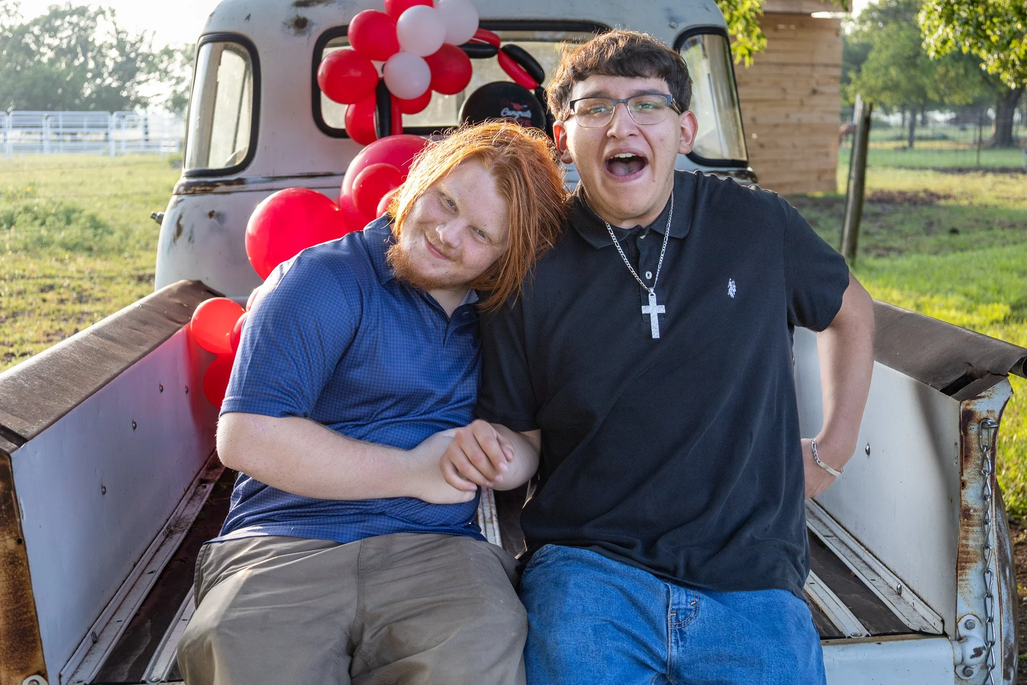 Two large young men sitting in the bed of a vintage pickup truck decorated with red and white balloons on a grassy field, with trees and a small wooden building in the background, during daytime.