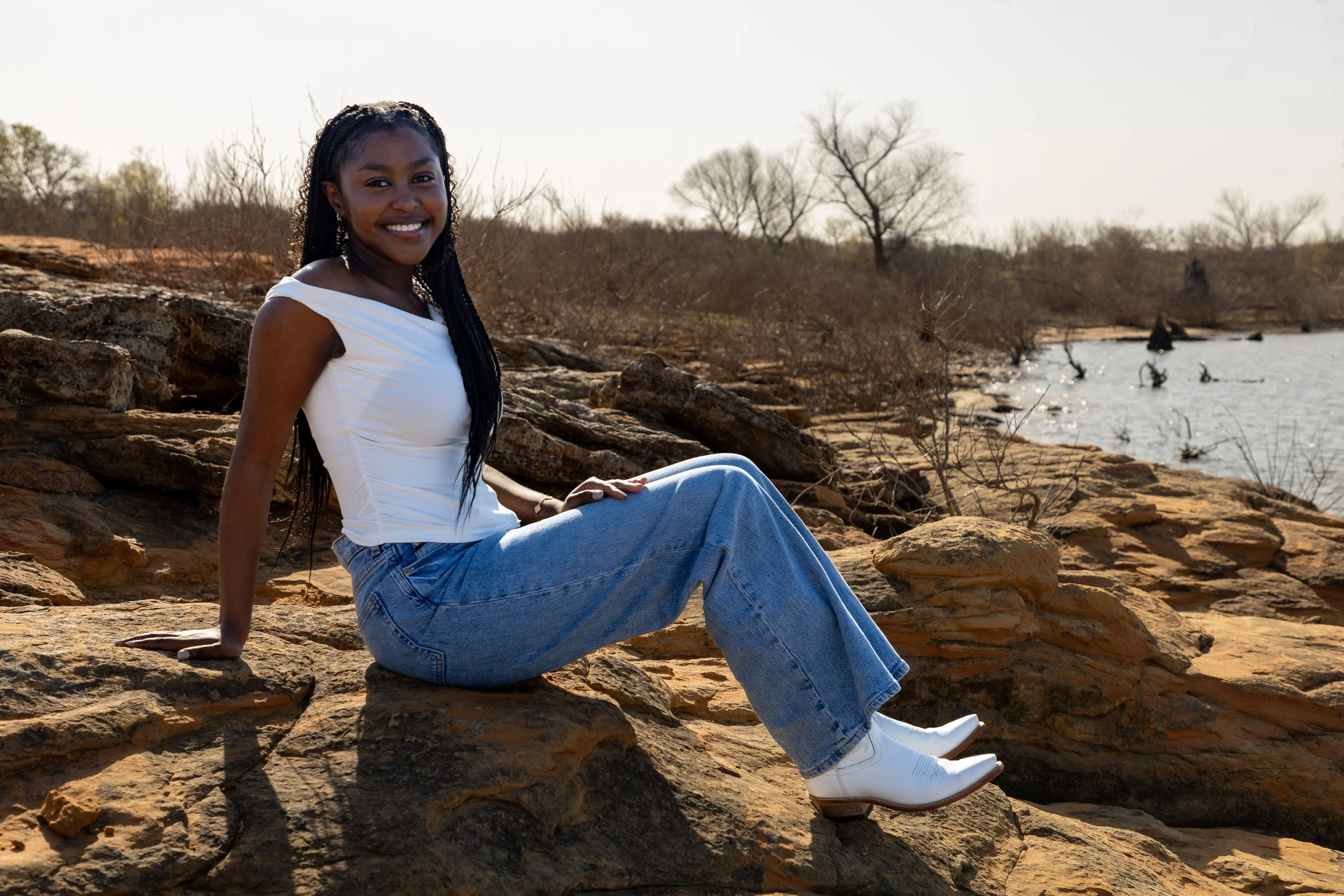 A young woman with long braided hair, wearing a white sleeveless top, blue jeans, and white cowboy boots, sitting on rocks by a river with leafless trees in the background.