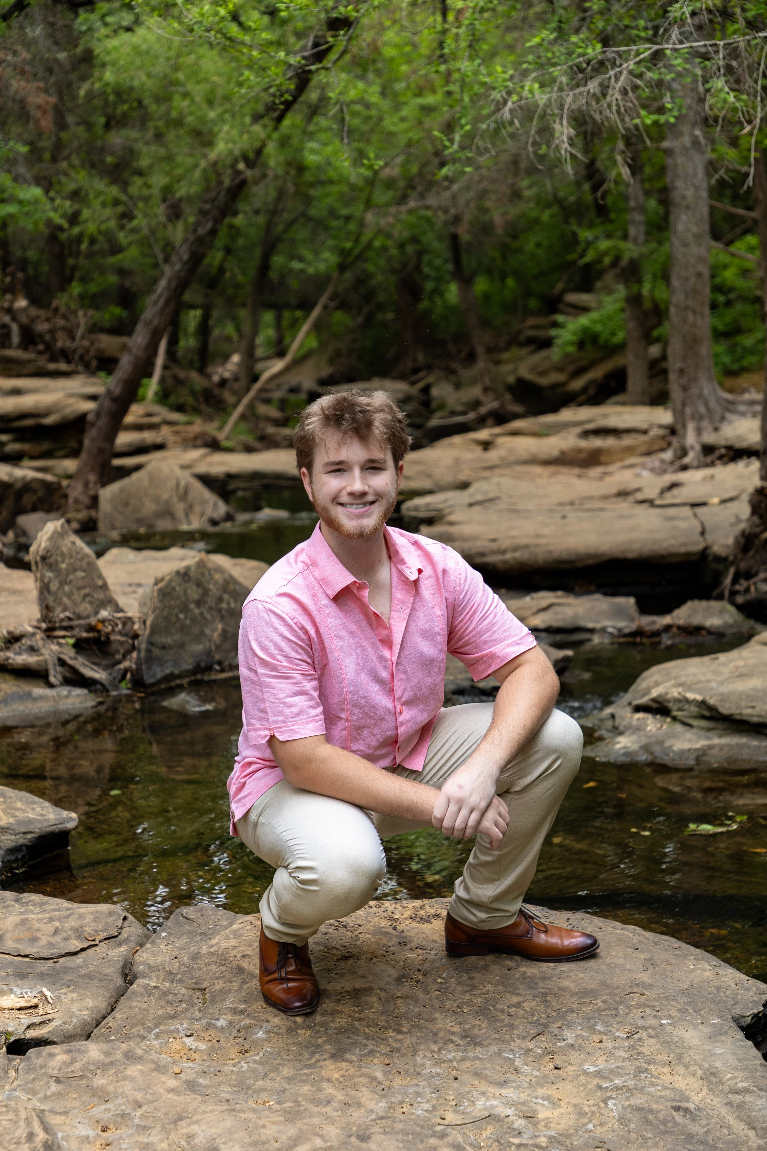 A young man with brown hair and a beard, wearing a pink short-sleeved button-down shirt, beige pants, and brown shoes, squatting on a large rock in a creek with a forest background.