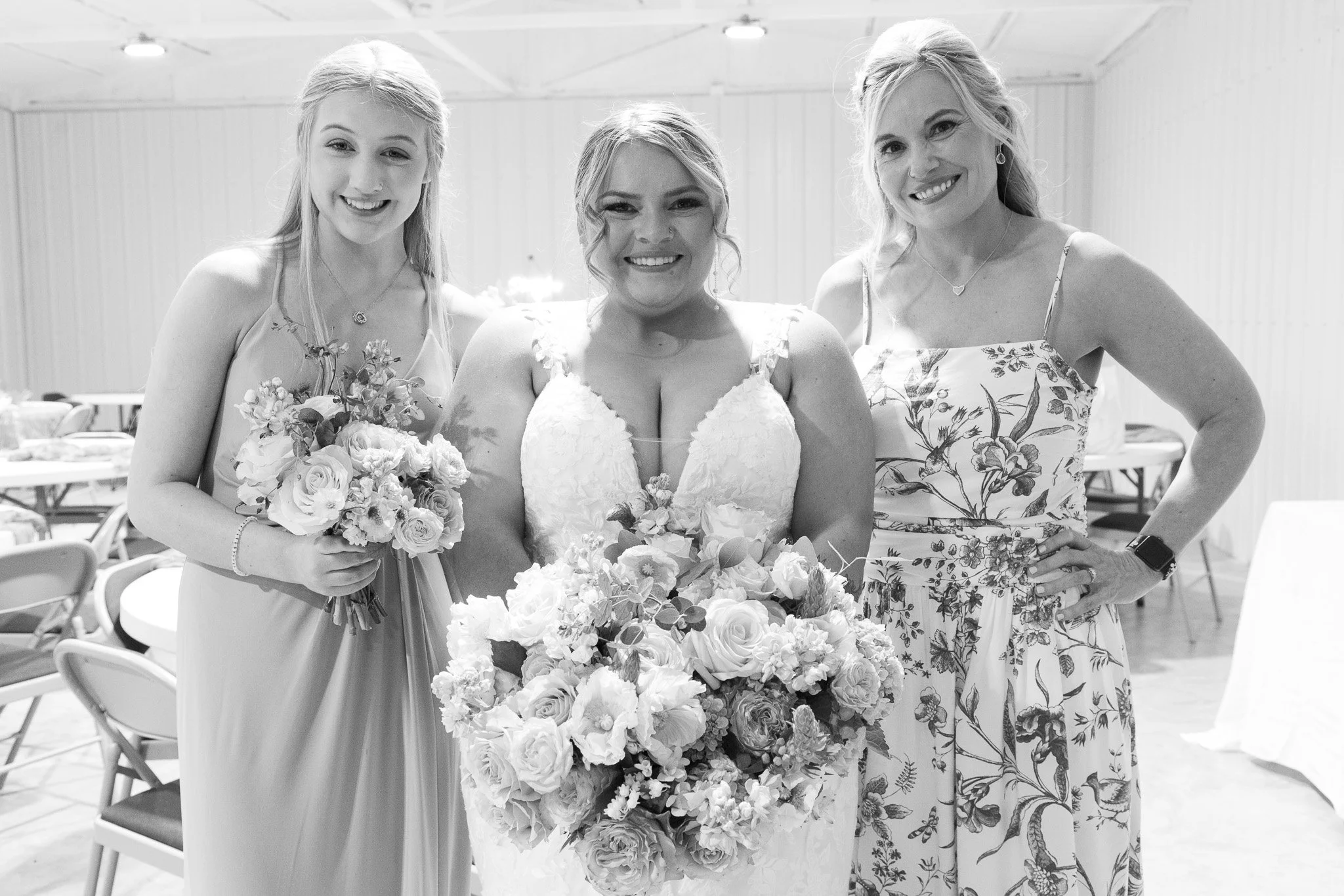 Three women, including a bride in a wedding dress holding a large bouquet, stand together indoors at a wedding reception. Two women are dressed in floral dresses, smiling and posing for the photo.