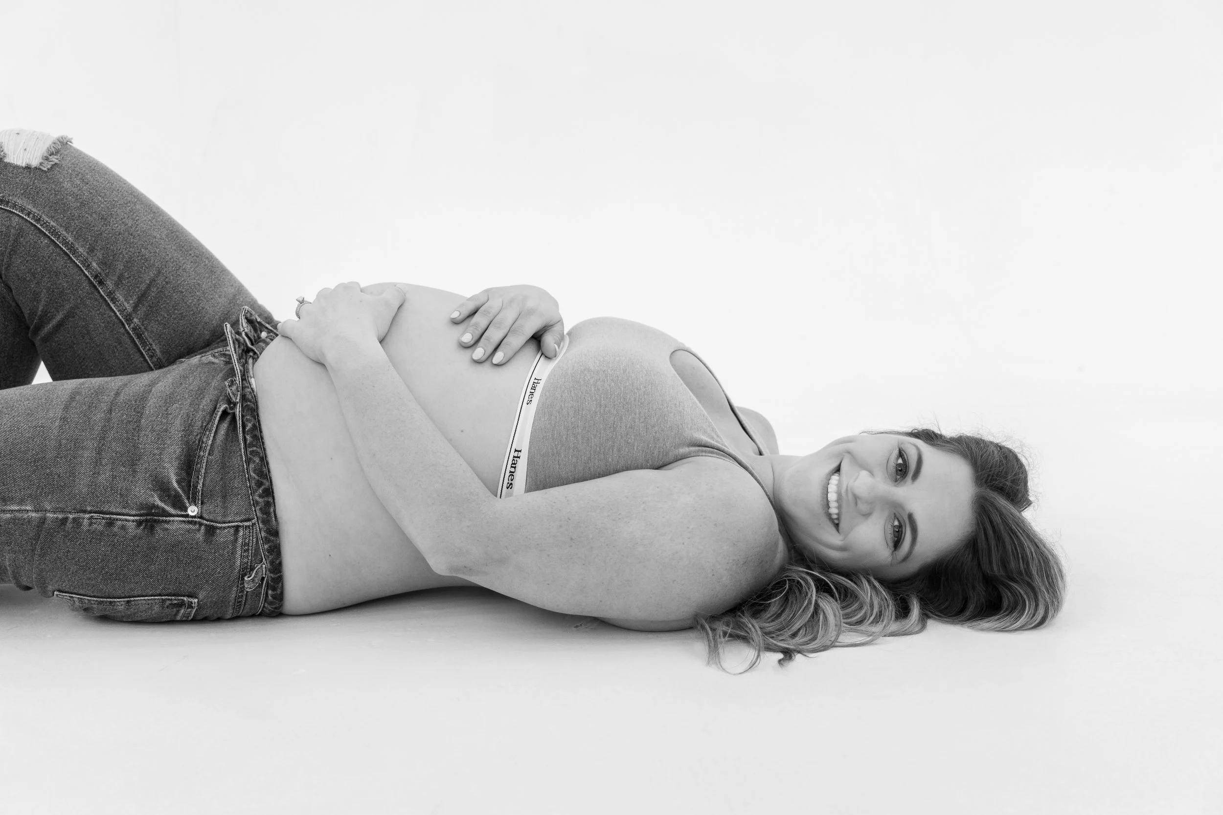 A woman lying down on her side, smiling, with her hands resting on her pregnant belly, wearing a tank top and jeans.