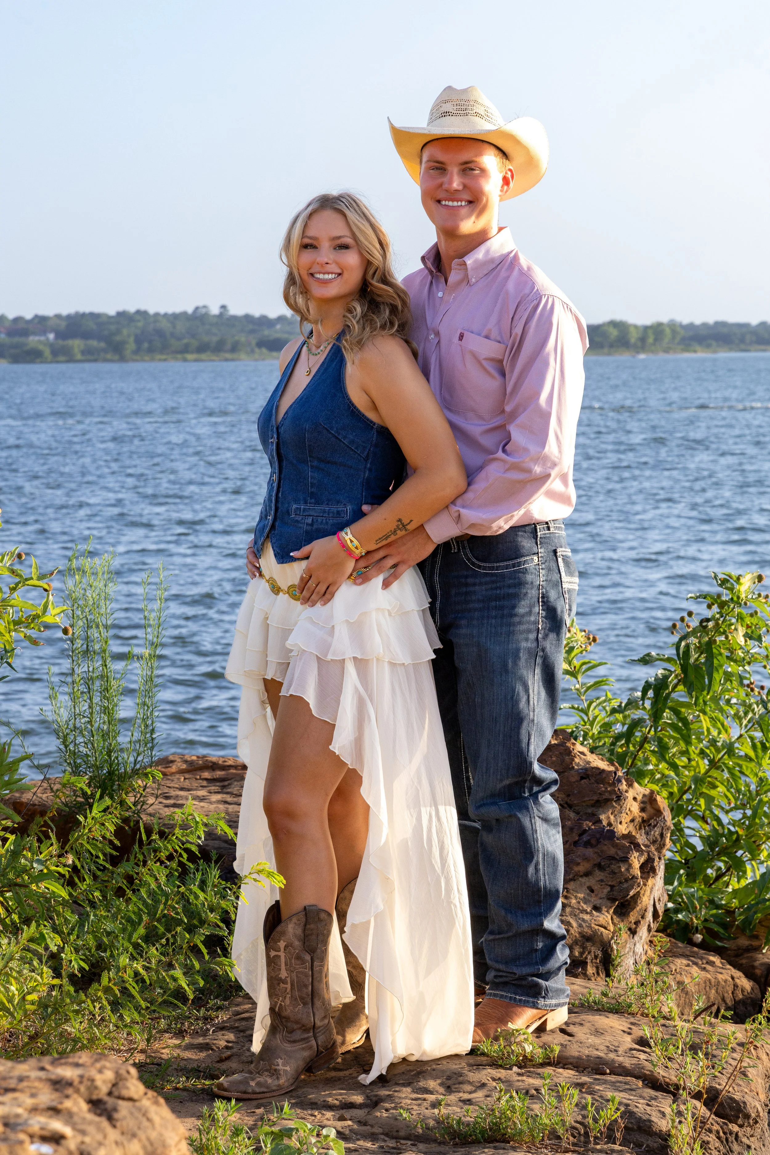 A smiling couple standing outdoors by a body of water, surrounded by rocks and green plants, with a distant tree line under a clear sky, dressed in casual cowboy attire.