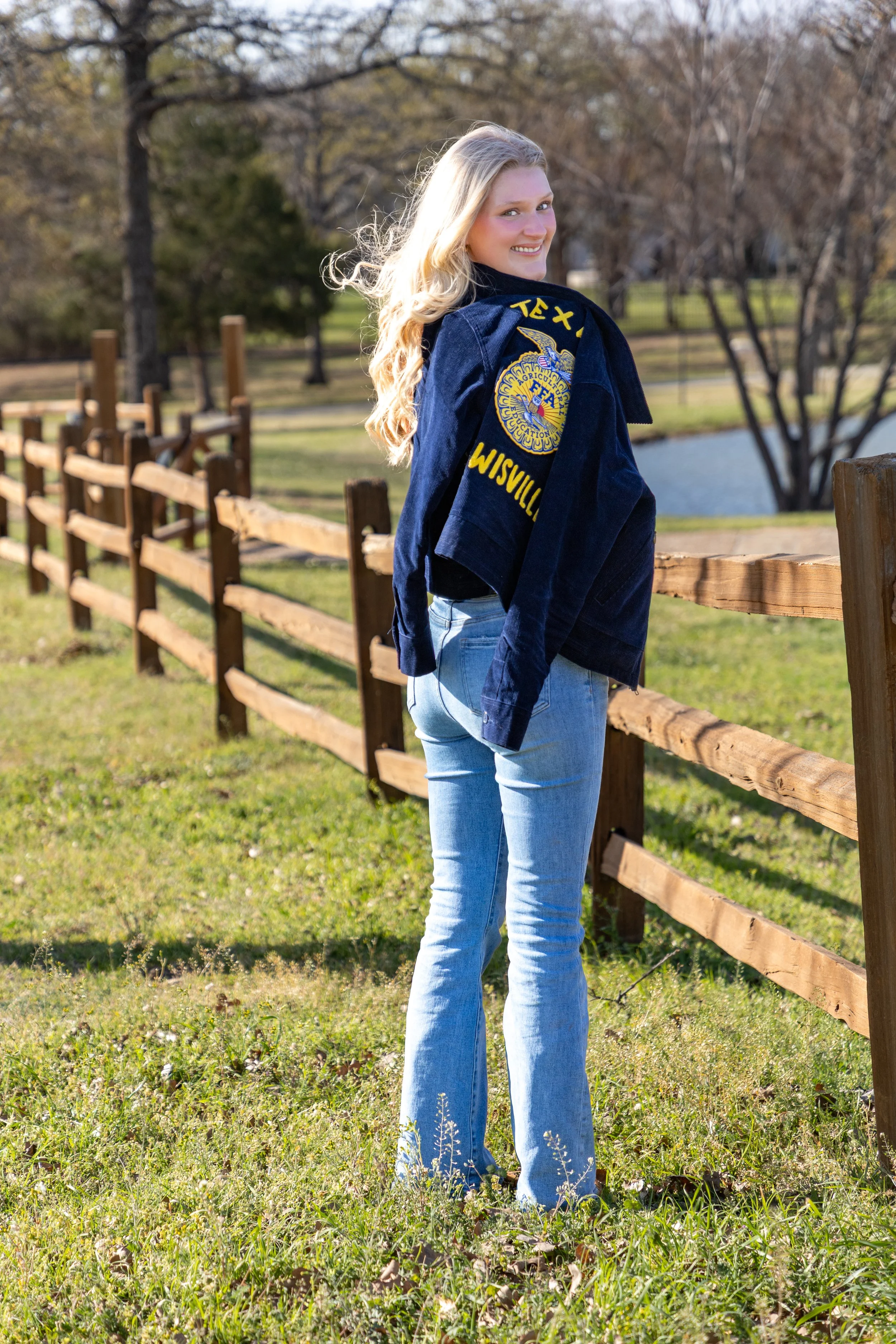 Young woman standing outdoors next to a wooden fence, smiling, wearing a navy blue quilted jacket with ffa emblem and yellow letters, jeans, and long blonde hair, in a park during autumn.