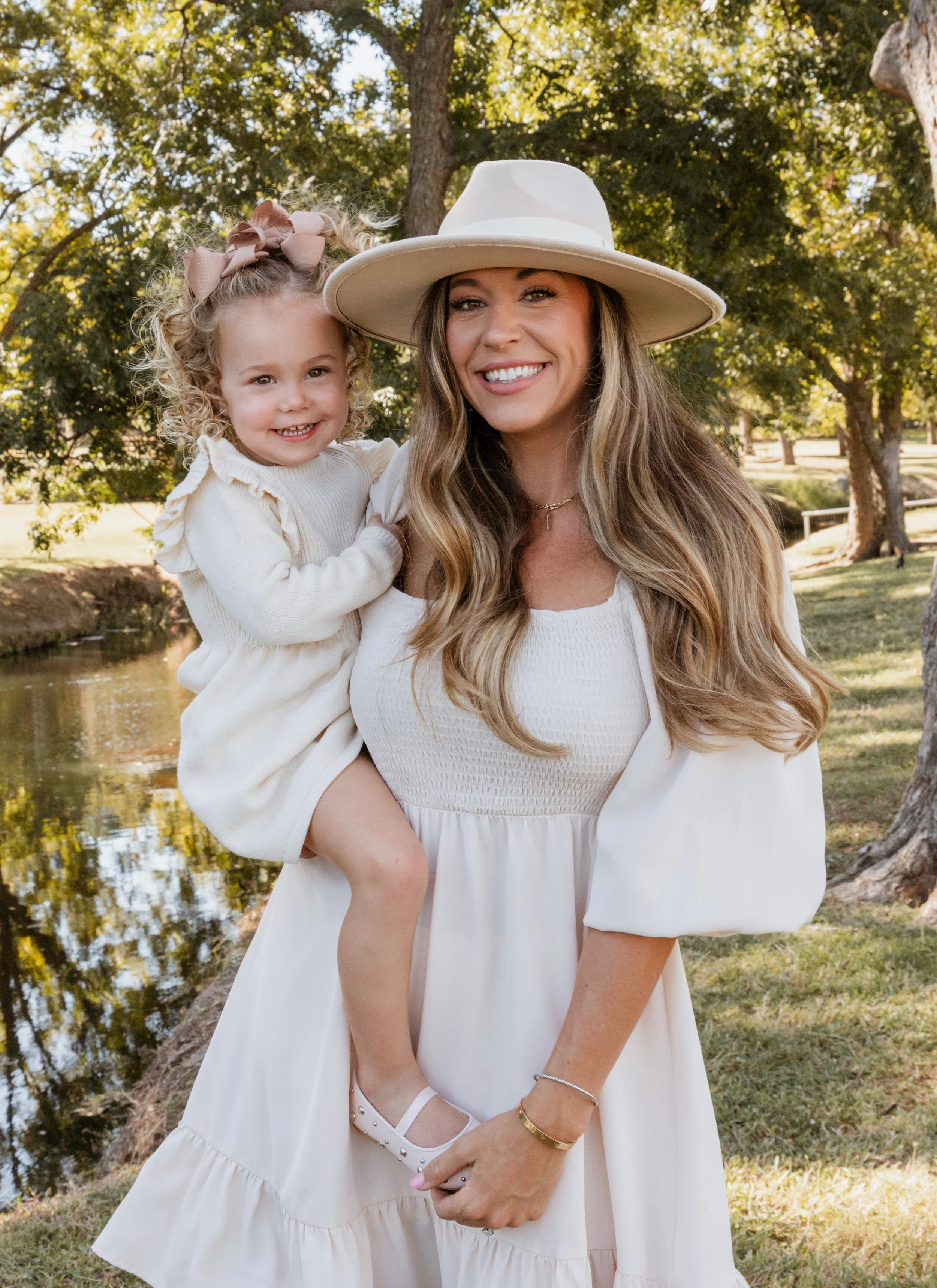 A woman smiling while holding a young girl on her shoulder outdoors in a park with trees and a pond in the background.