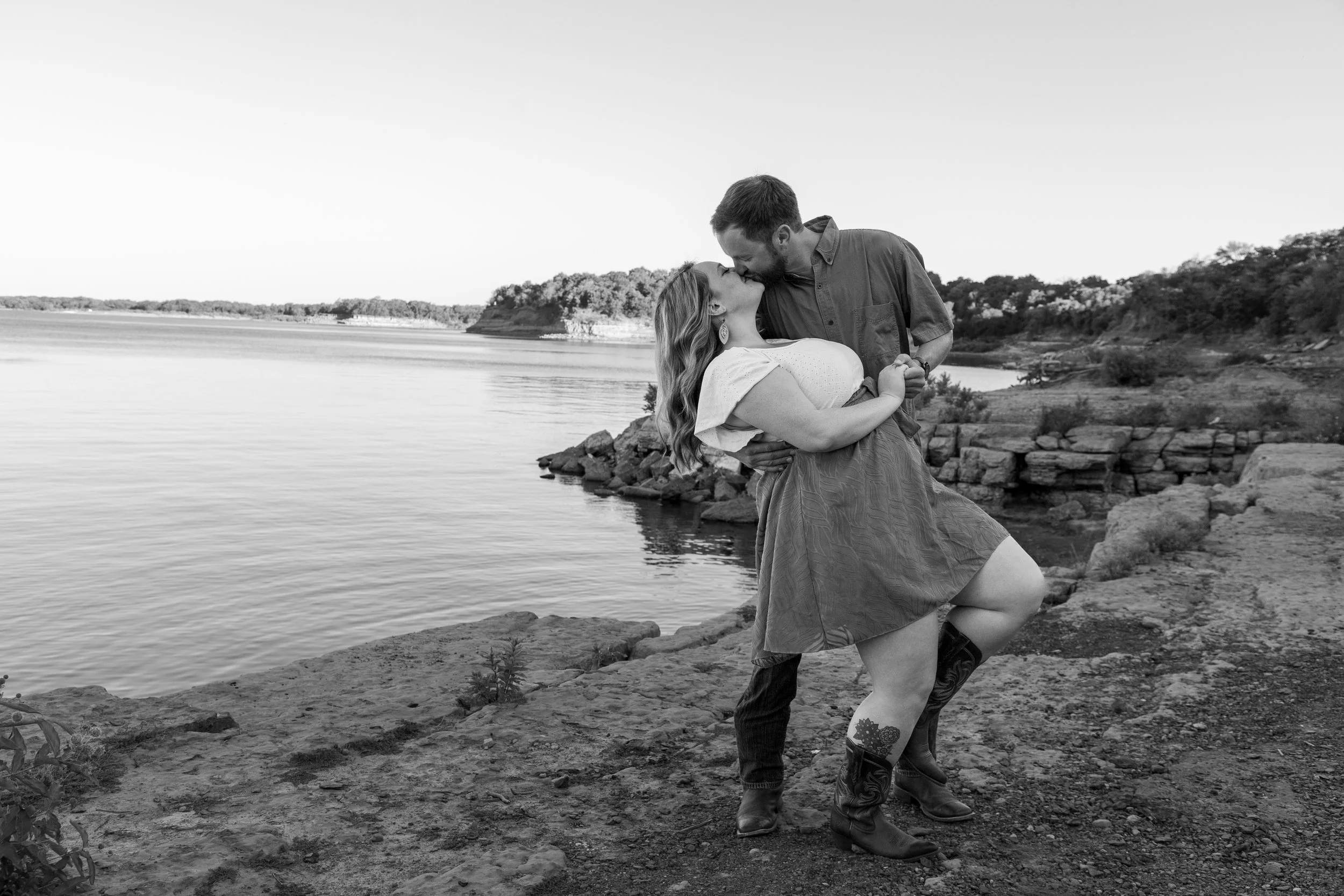 A couple sharing a kiss on a rocky riverside, with the woman leaning back and the man holding her, black and white photo.