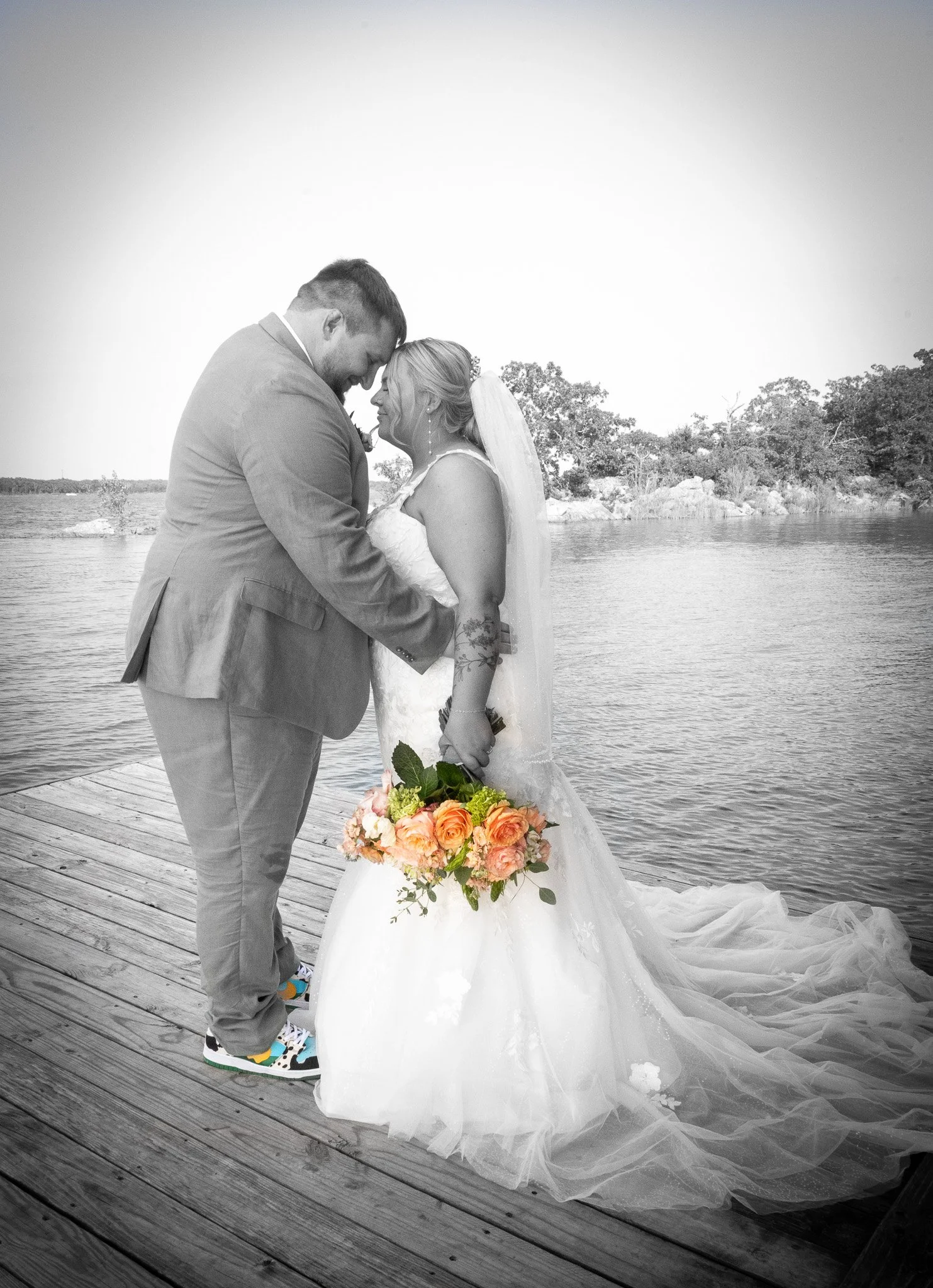 A bride and groom stand close together on a wooden dock by a body of water, smiling and holding hands, with the bride holding a bouquet of pink and orange roses. The photo is primarily black and white, with only the flowers in color.