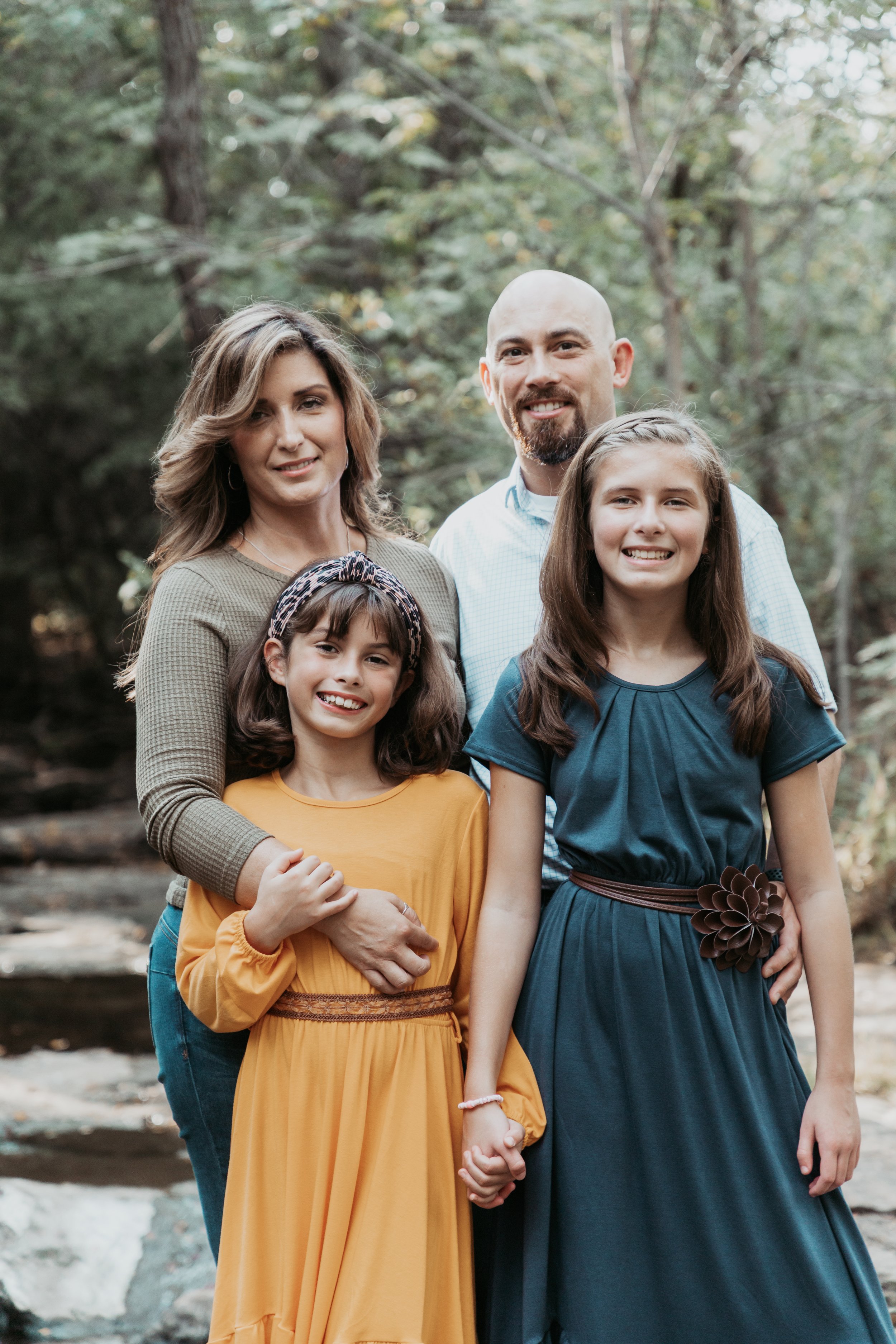 A family of five posing outdoors in a wooded area, smiling at the camera.