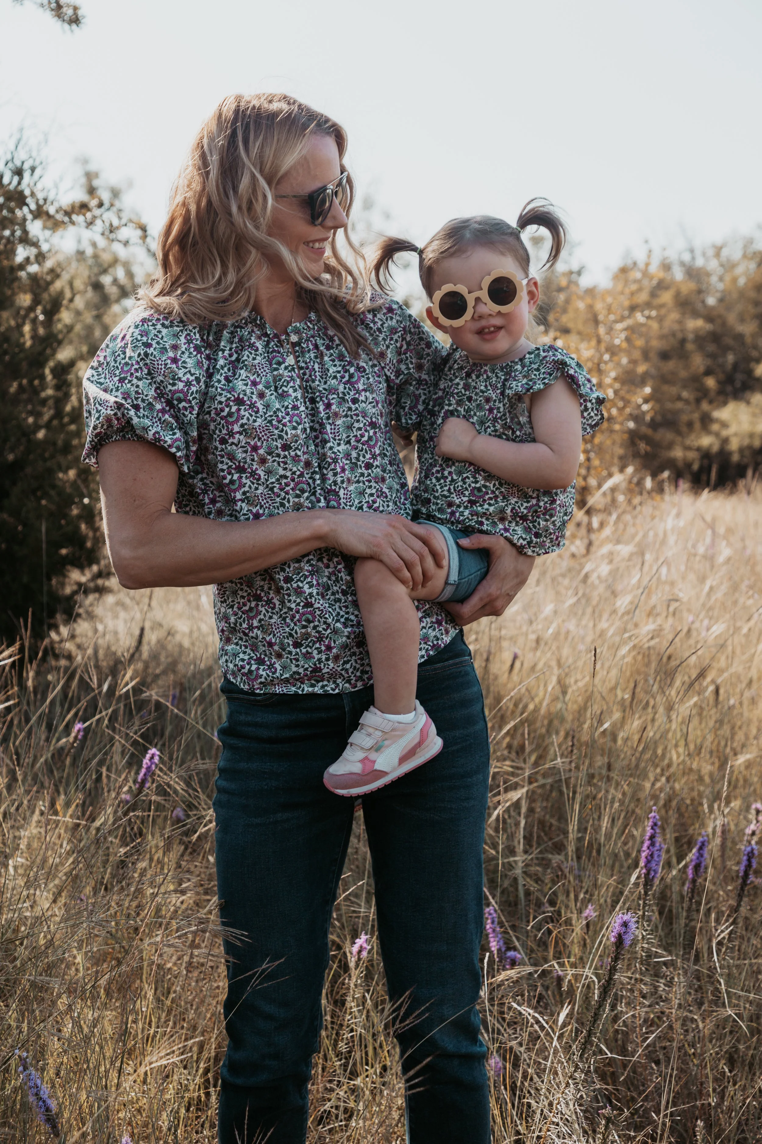 A woman holding a young girl in a field of tall grass and purple flowers on a sunny day.