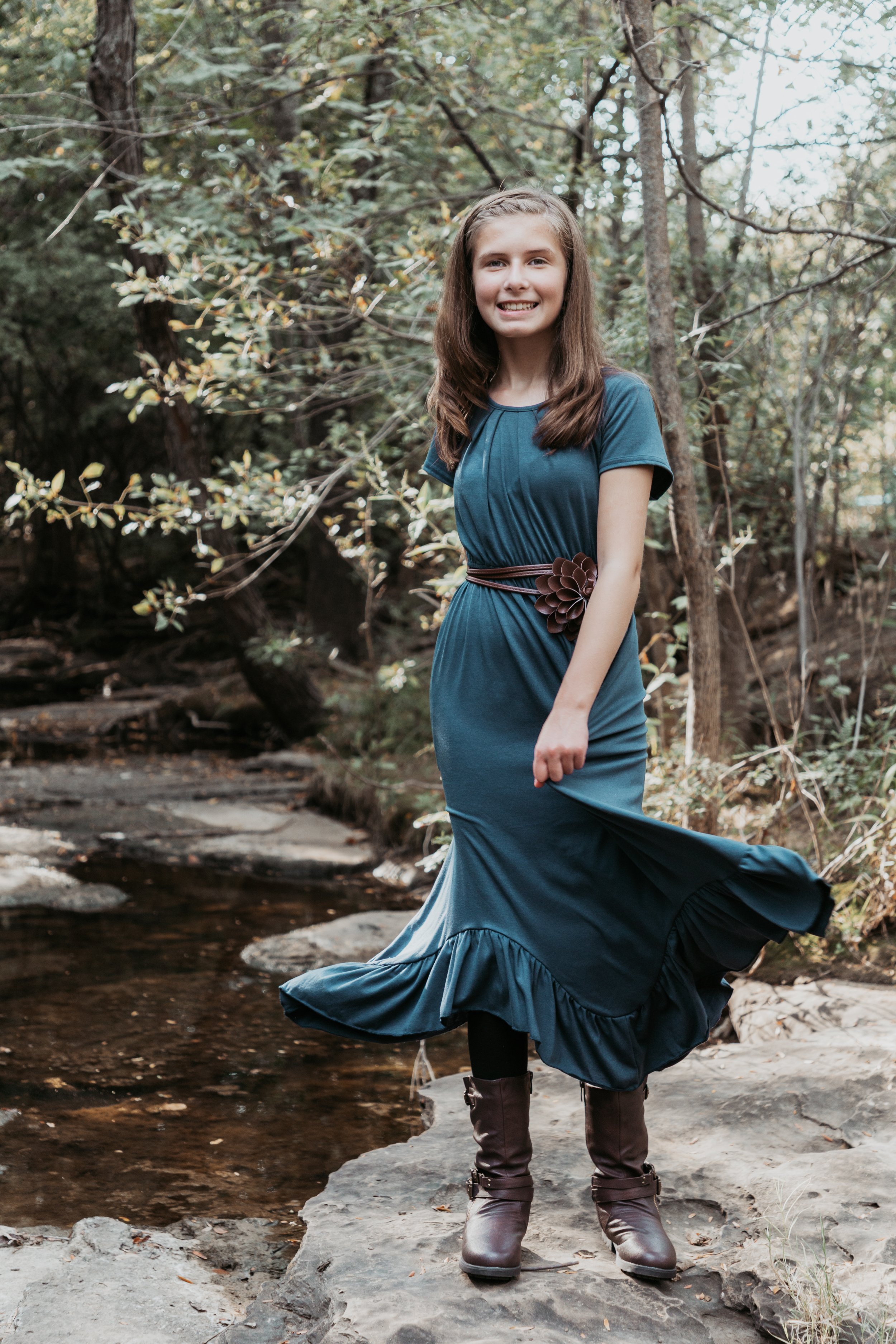 A young girl in a teal dress with a ruffled hem, brown belt, and brown boots stands on a rocky surface near a small creek in a wooded area, smiling at the camera.