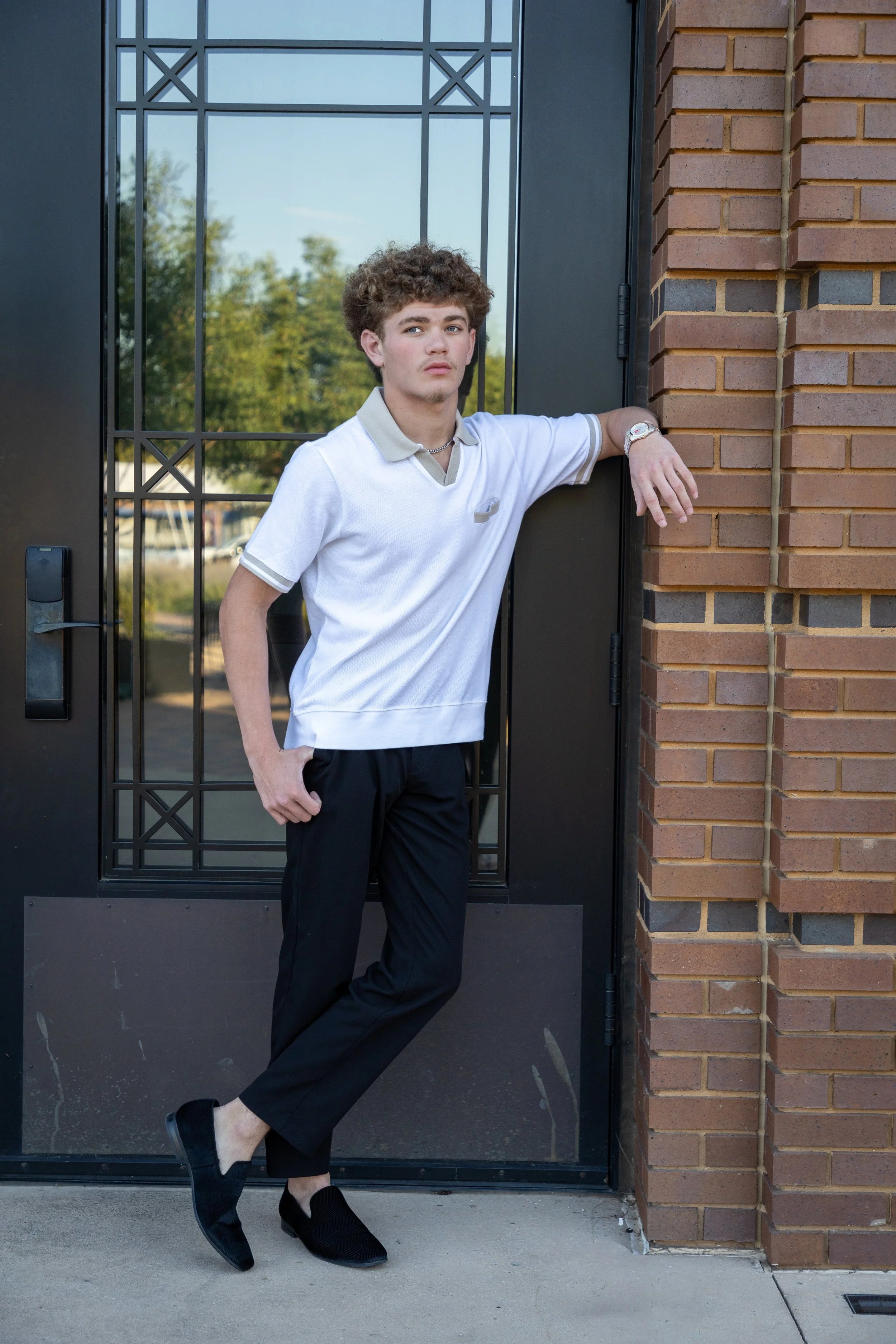 A young man with curly hair leaning against a brick wall beside a glass door, wearing a white polo shirt, black pants, and black slip-on shoes.